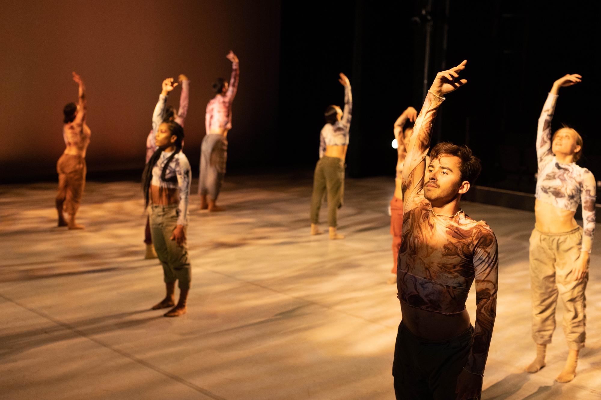 eight dancers stand on stage with their right arms raised, washed in warm, golden stage lighting