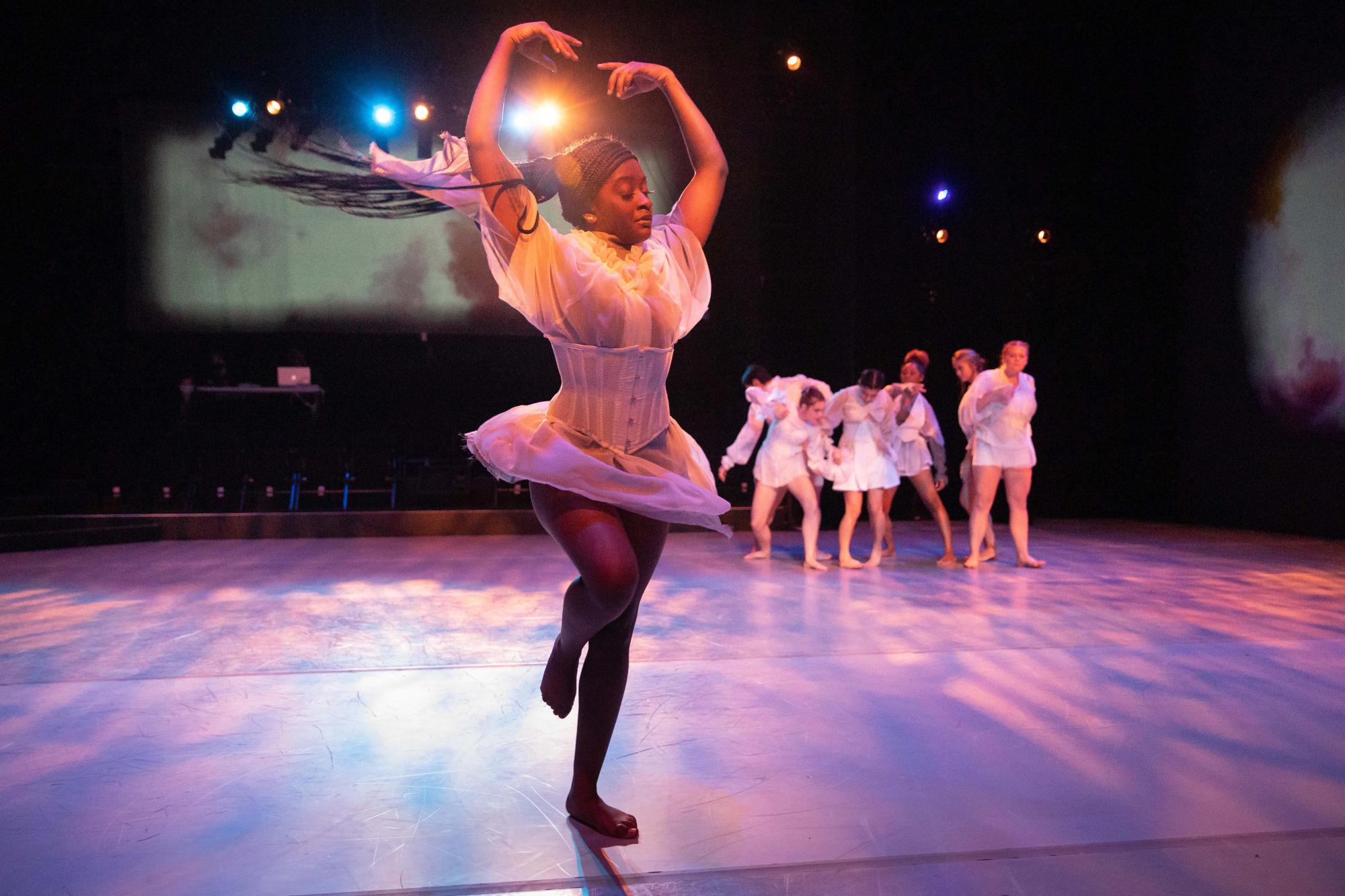 A dancer in all white pirouettes, with a group of dancers behind her