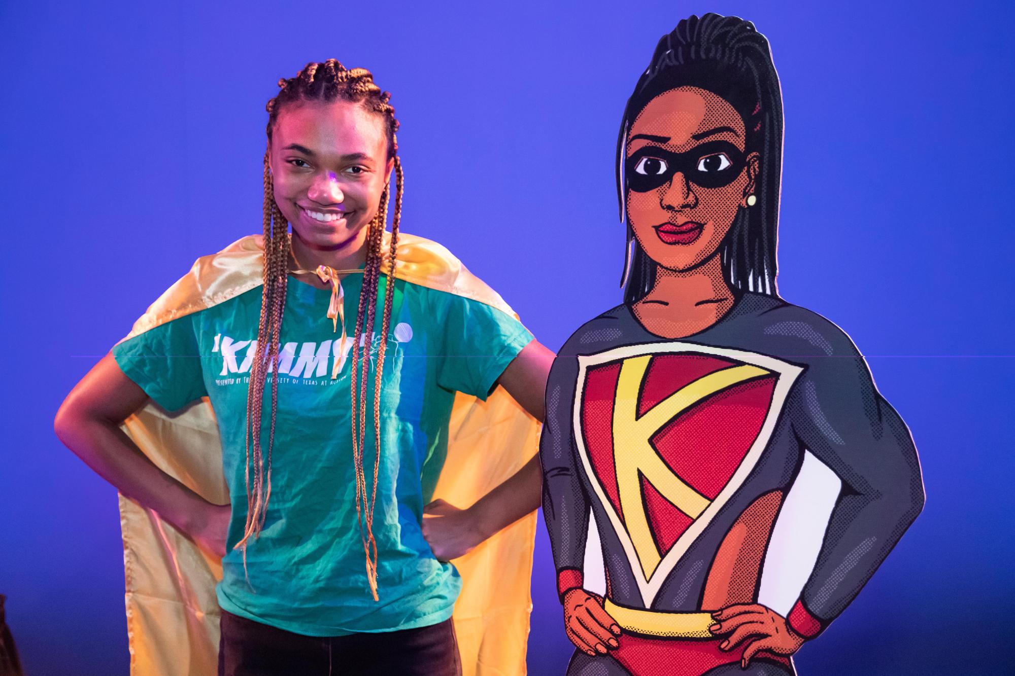 an actress with a yellow cape and braids poses next to a cardboard cutout of the superhero version of "Kemi," the character she voiced