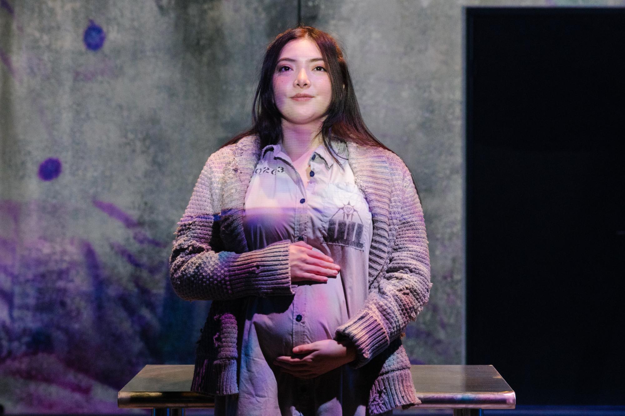 actress with her hands on her baby bump looks confidently out into the audience