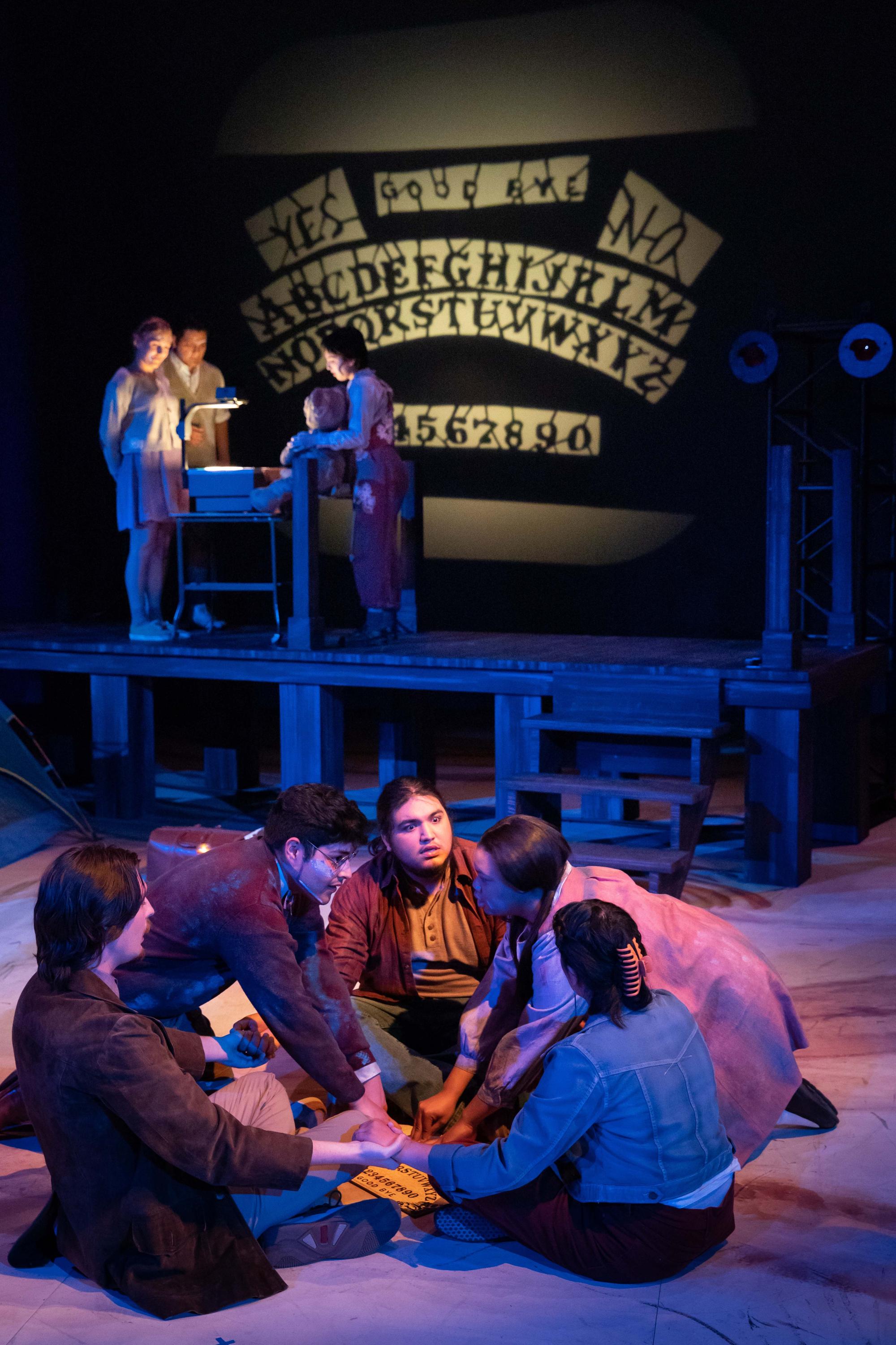 three people sit around an ouija board while five "ghost children" manipulate what it says