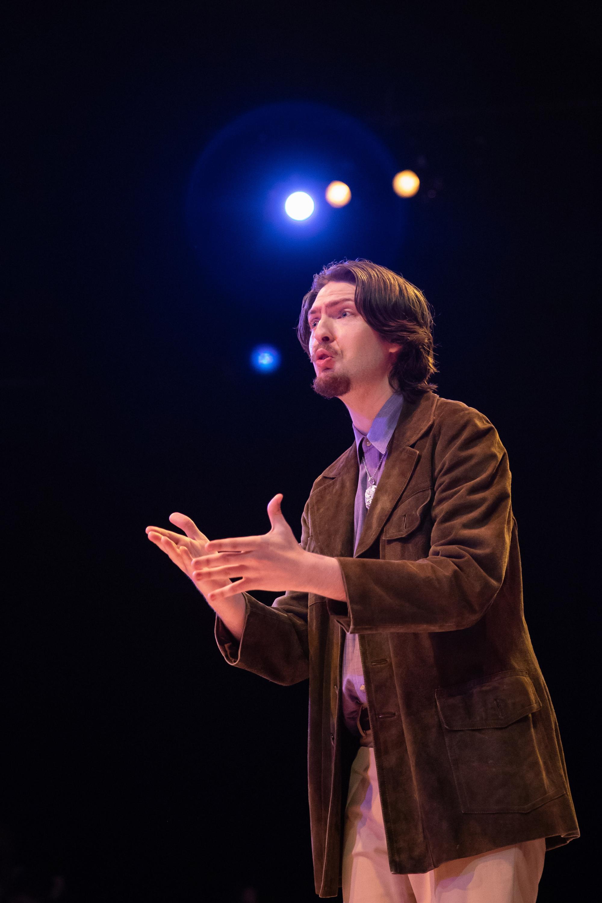 a man in a corduroy jacket speaks, holding his hands out, with stage lights shining behind him 