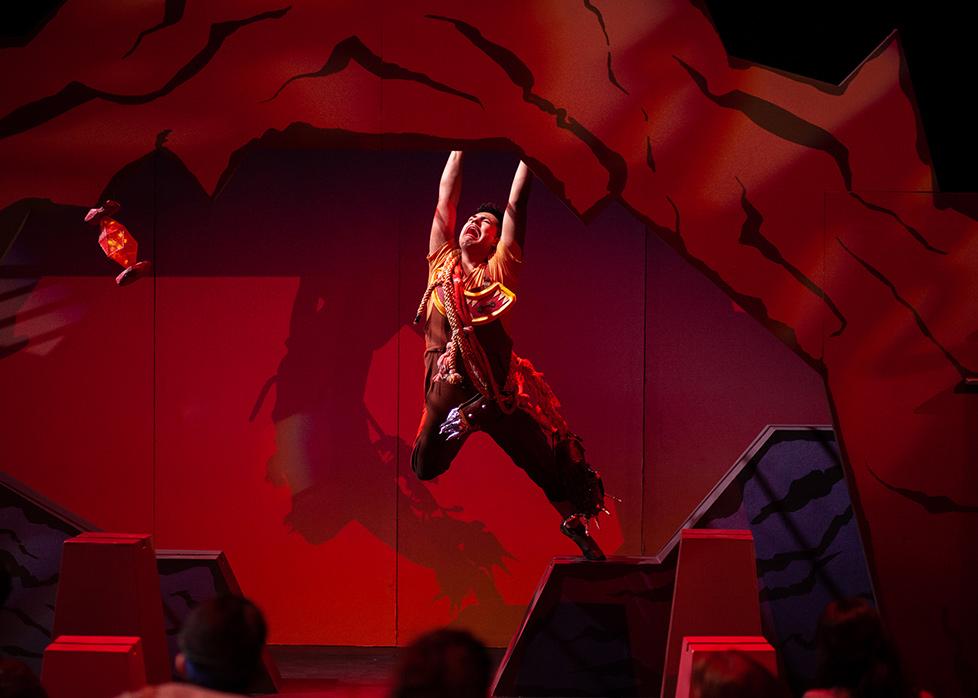 an actress hangs from a set piece against a red-lit backdrop