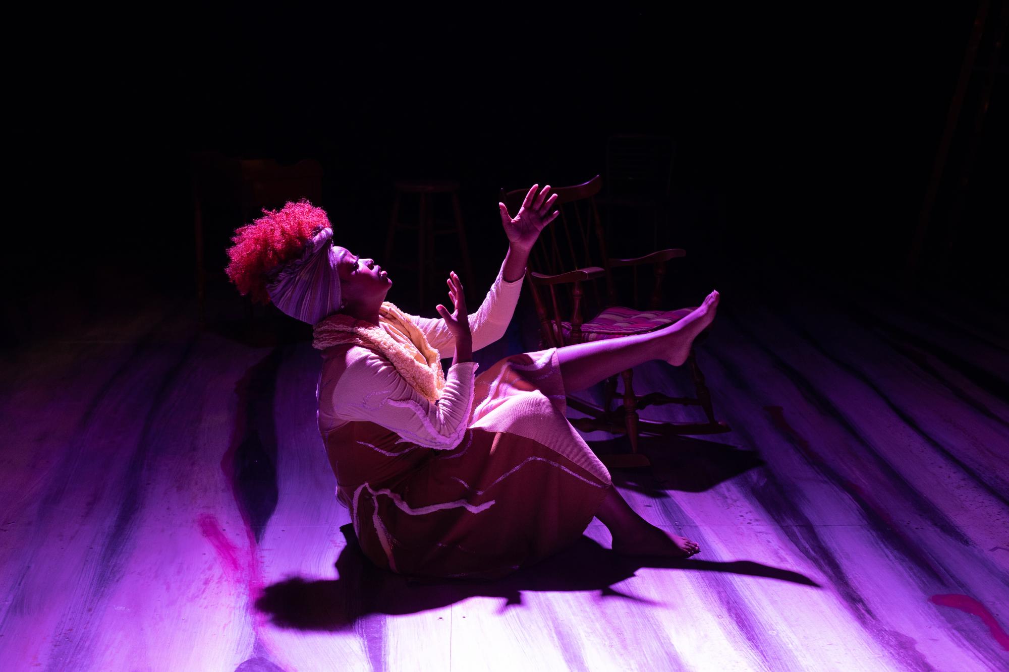 woman sits on the ground with her arms reaching upward, washed in purple stage light