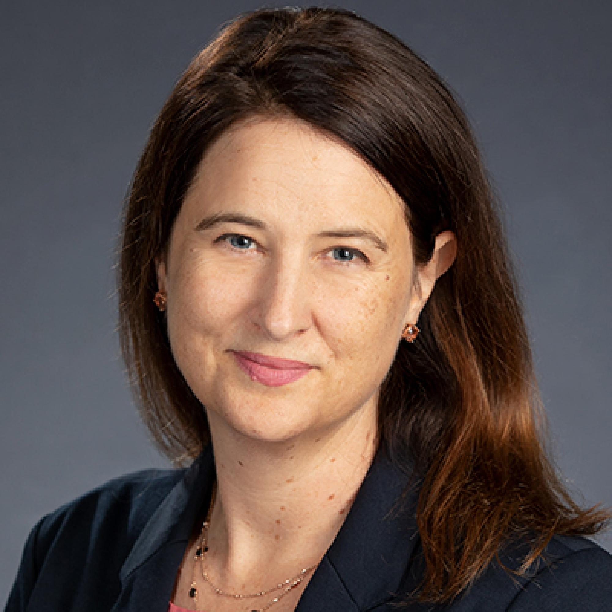 woman with long brown hair smiles, wearing a necklace and a black blazer