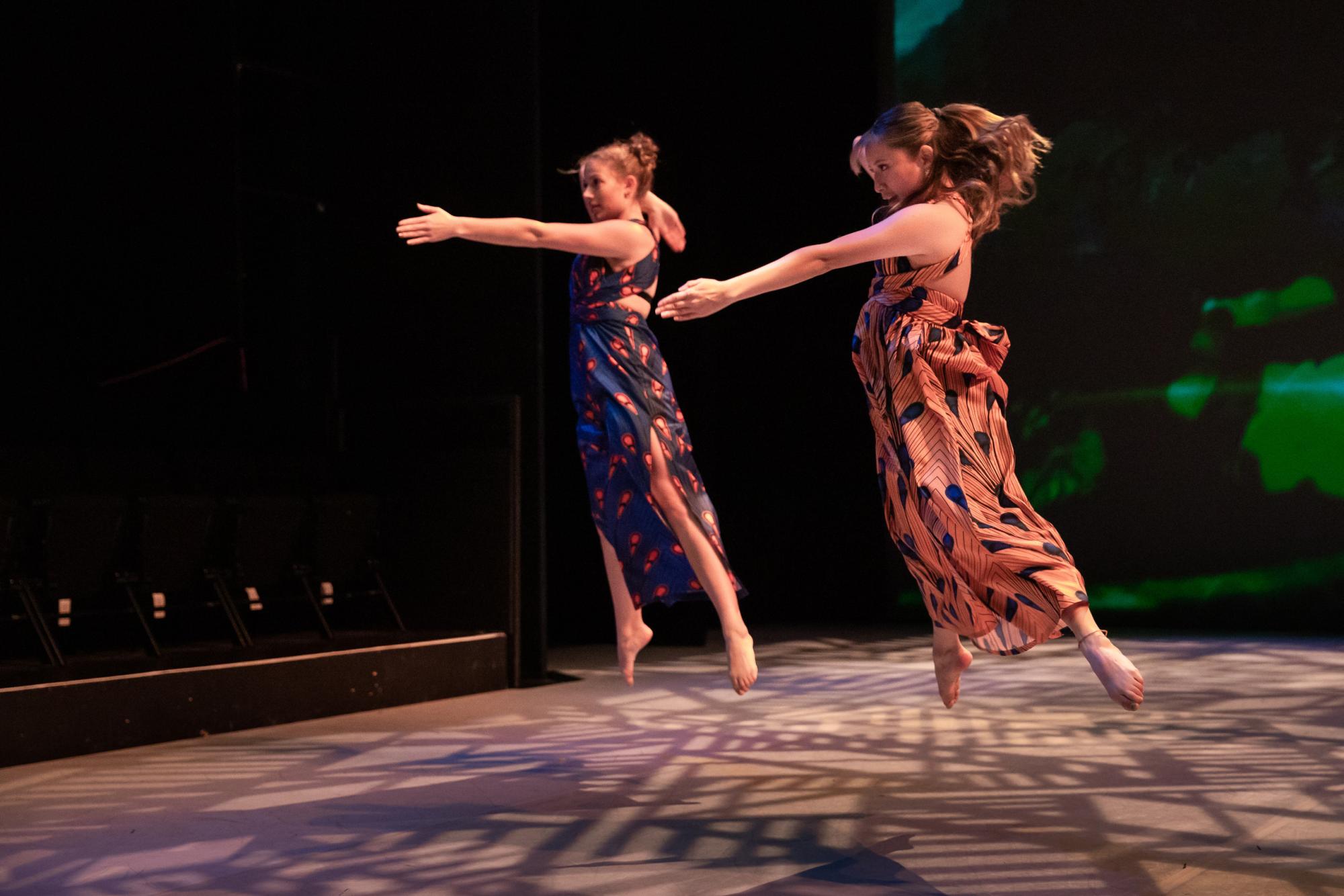 Two dancers dressed in animal print dresses jump in the air, with one arm outstretched 