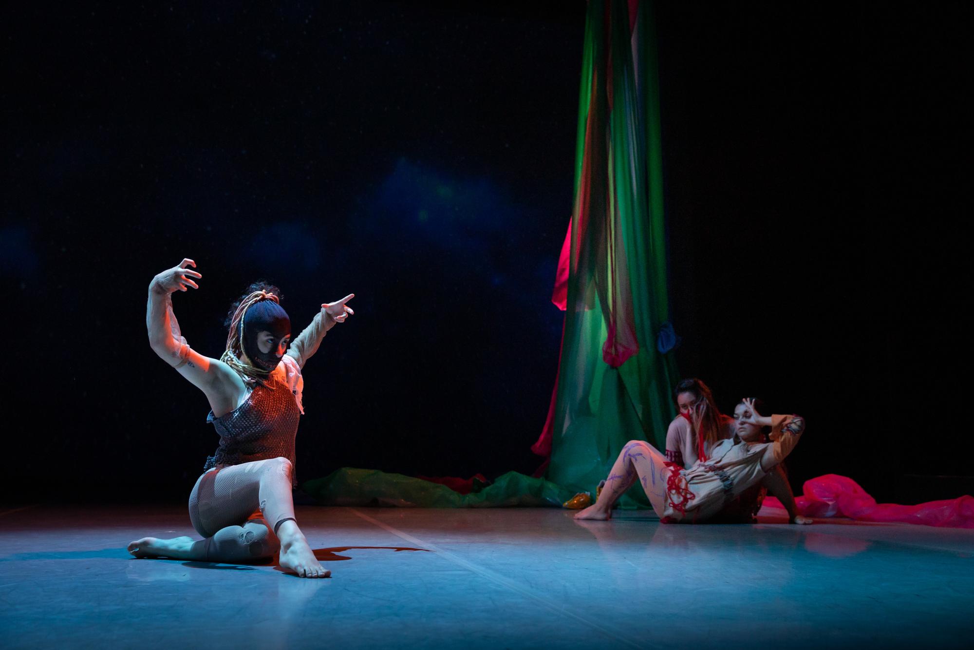 A dancer sits, arms stretched overhead, wearing a black balaclava 