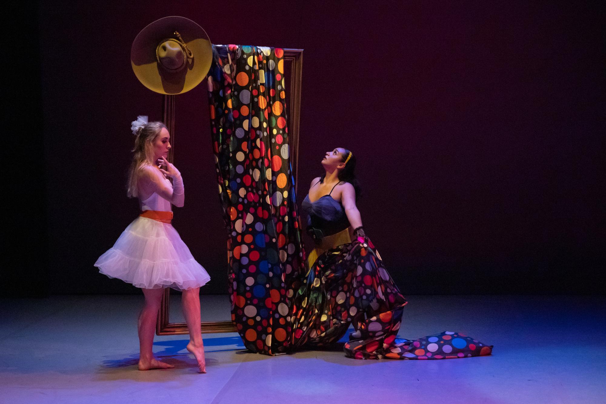 Two dancers stand on stage, one wrapped in a multicolored polkadot cloth