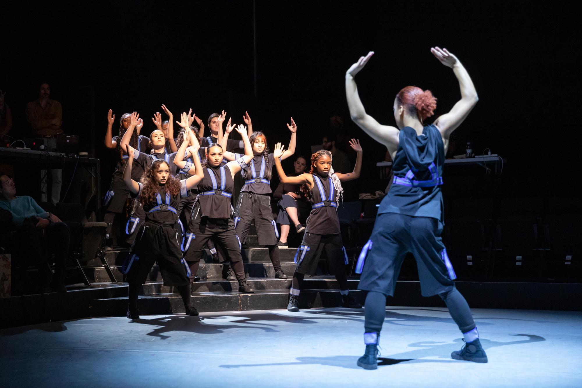 A group of dancers in technical garb pose with their arms above their heads, facing a single dancer who poses in the same way