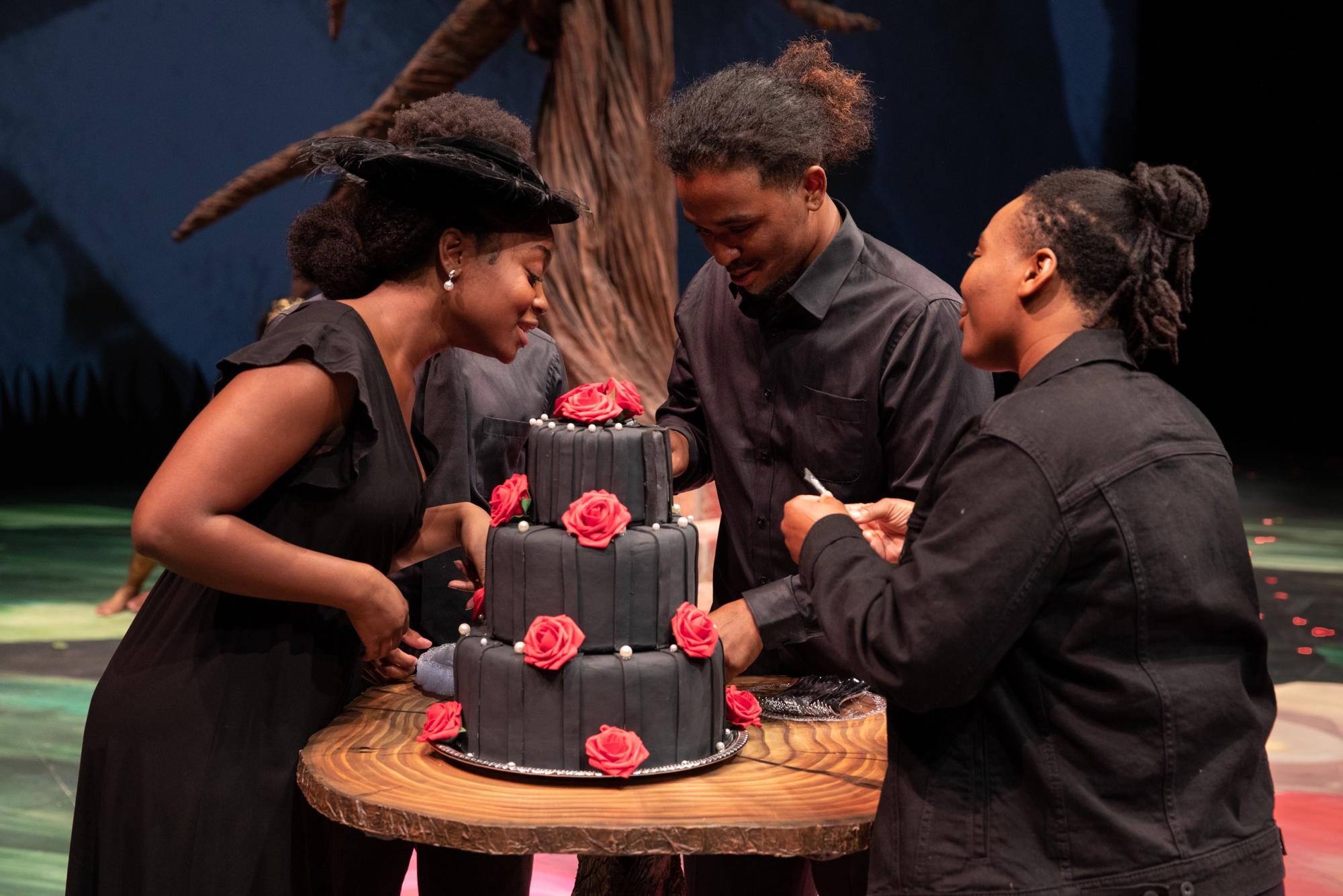 Three people dressed in black cut a black cake with red roses on it