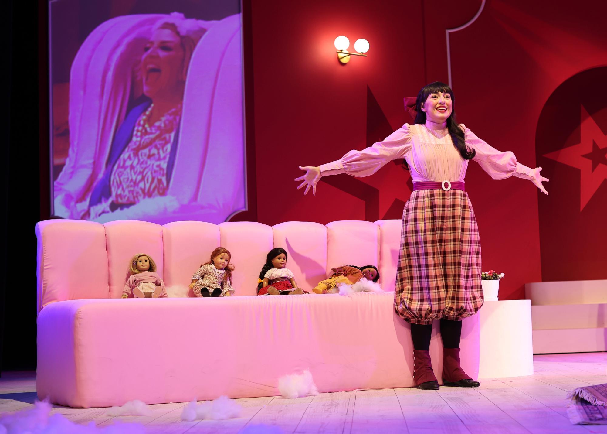 A woman spreads her arms out as 4 American girl dolls lie on a couch behind her