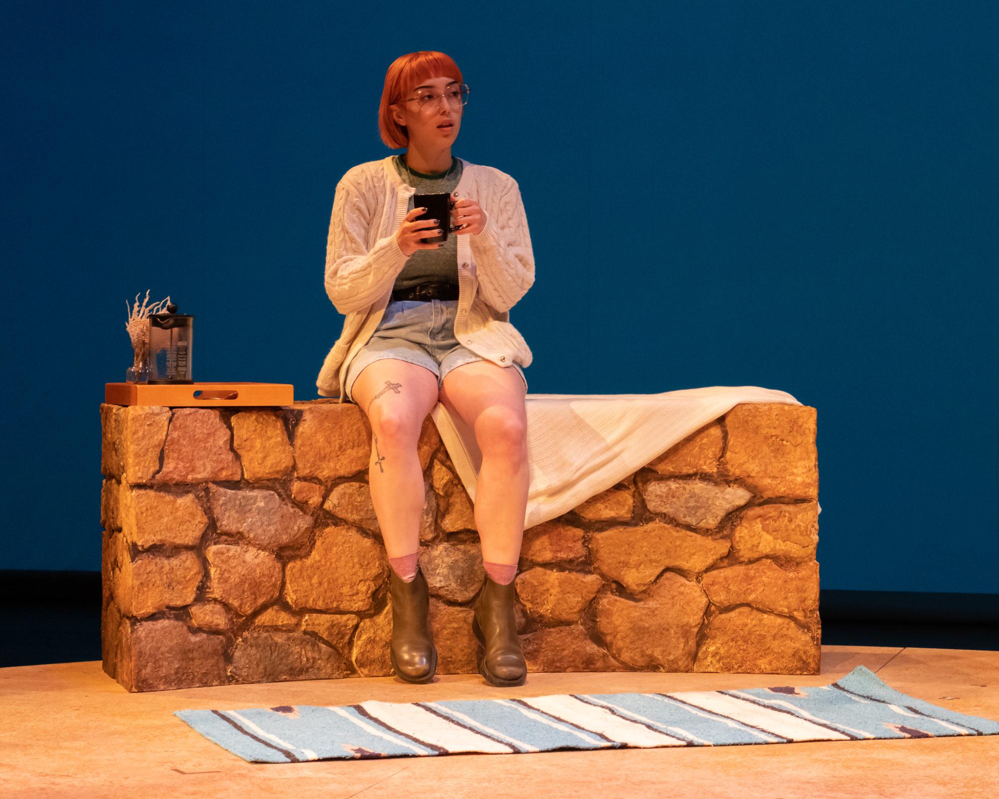 A person sits on a desert wall, nursing a cup of tea