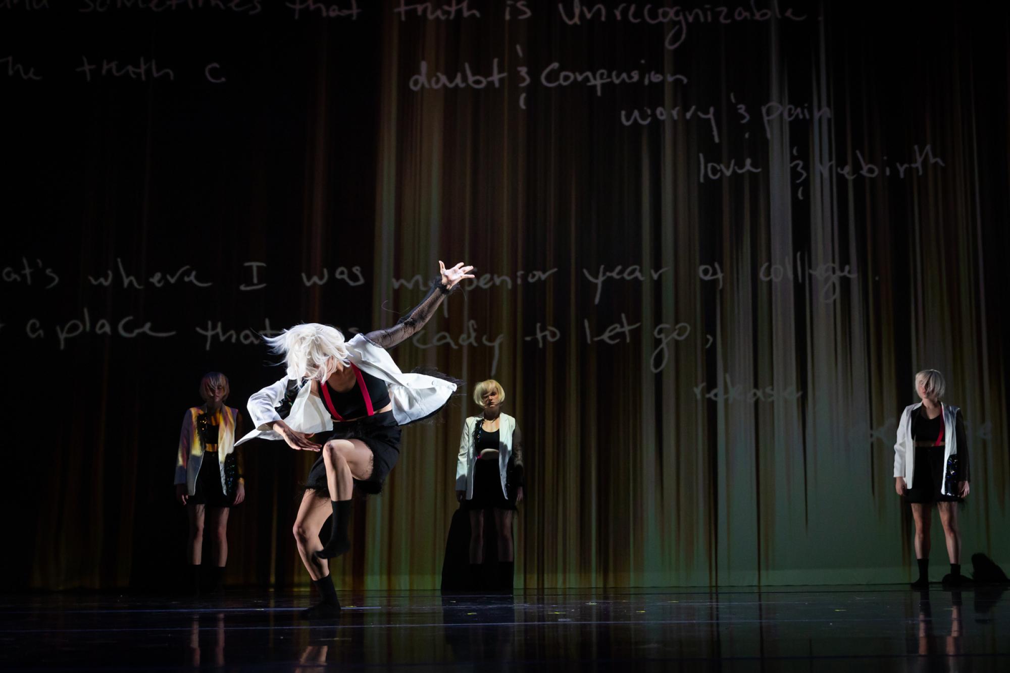 A dancer in a white wig poses as three other wigged dancers look on in the background