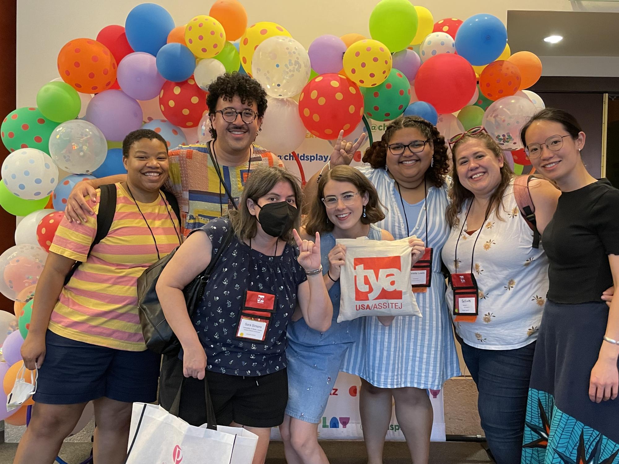 Faculty member Sara Simons and six UT grad students pose together in front of a colorful balloon arch at TYA/USA