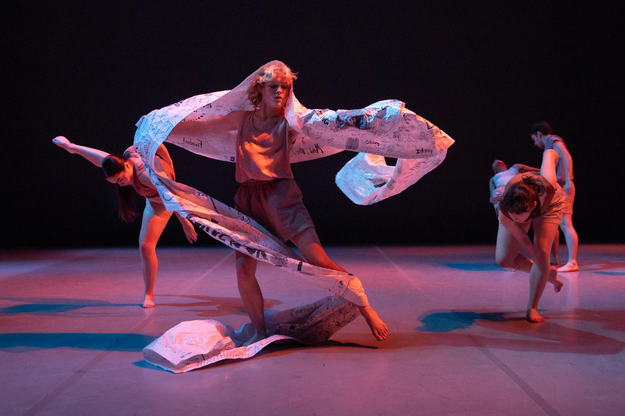 A dancer holds their arms out and their left leg slightly off the ground, with a large strip of paper draped around them