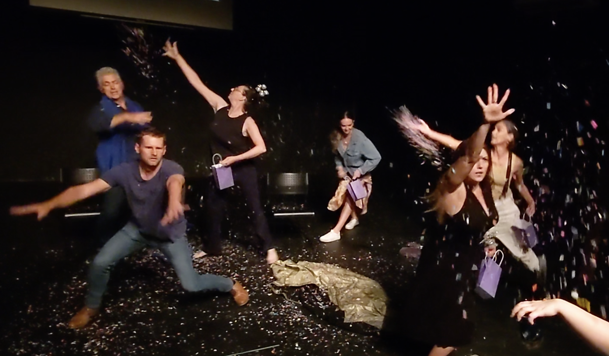 six performers move jovially through a dark performance space, throwing confetti all over the room