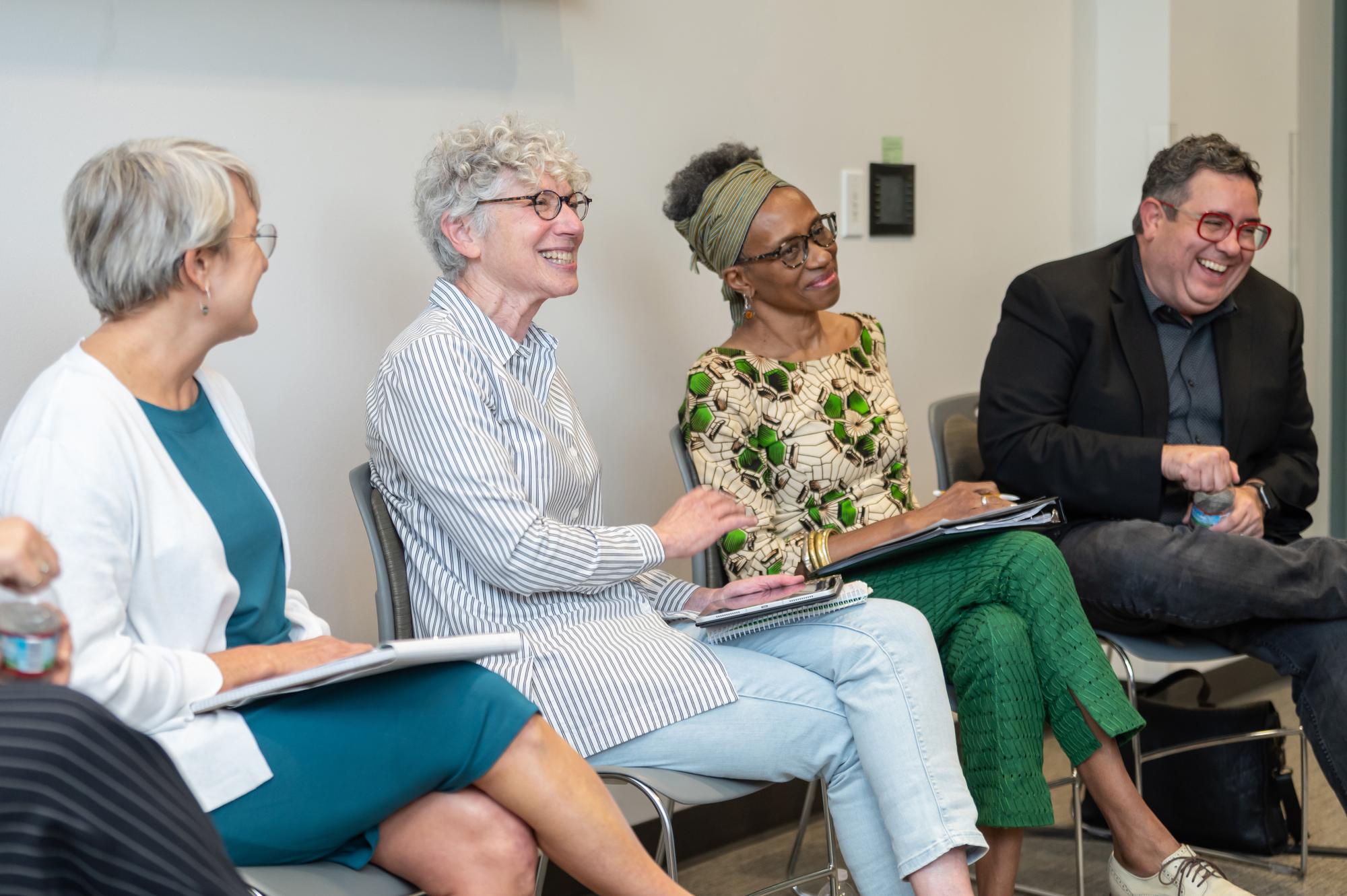 four faculty and former faculty smile and converse while sitting on chairs as part of a panel discussion