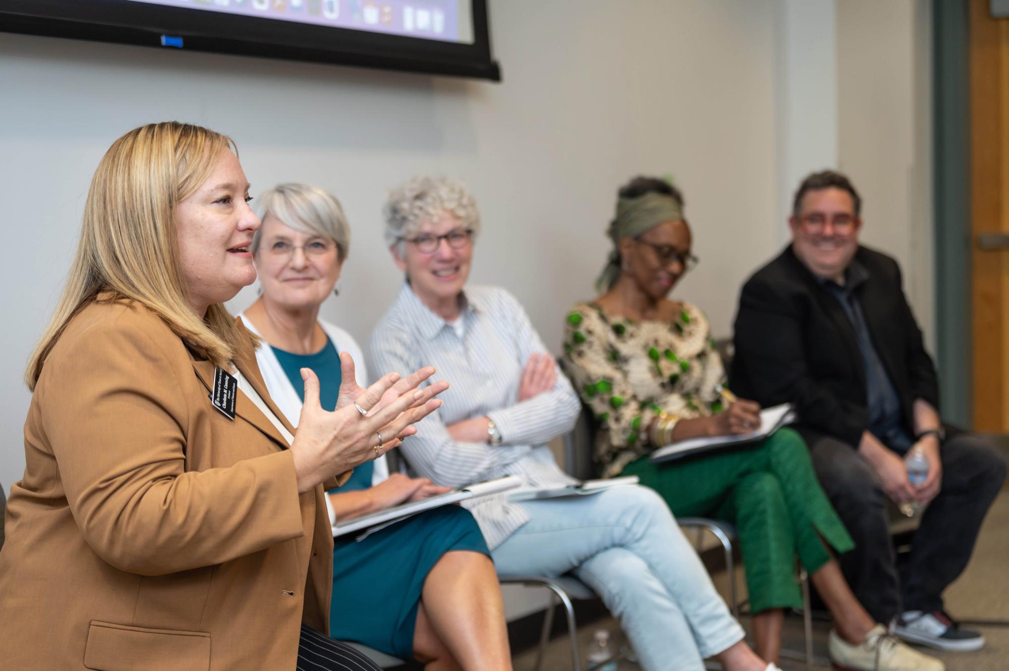 Five panelists sit at the front of the classroom. One is in the foreground speaking with her hands in front of her, while four other panelists watch in the background