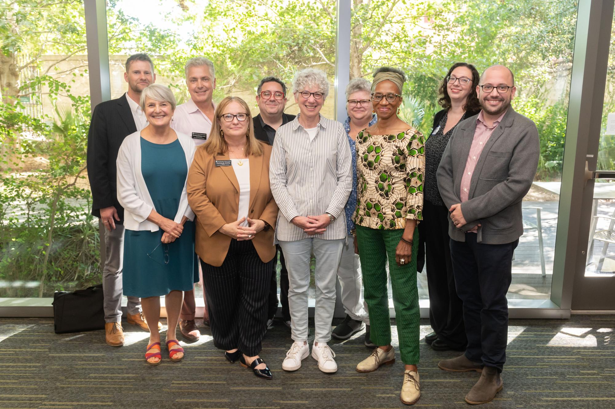 10 faculty and former faculty smile and stand in front of large windows with trees behind them