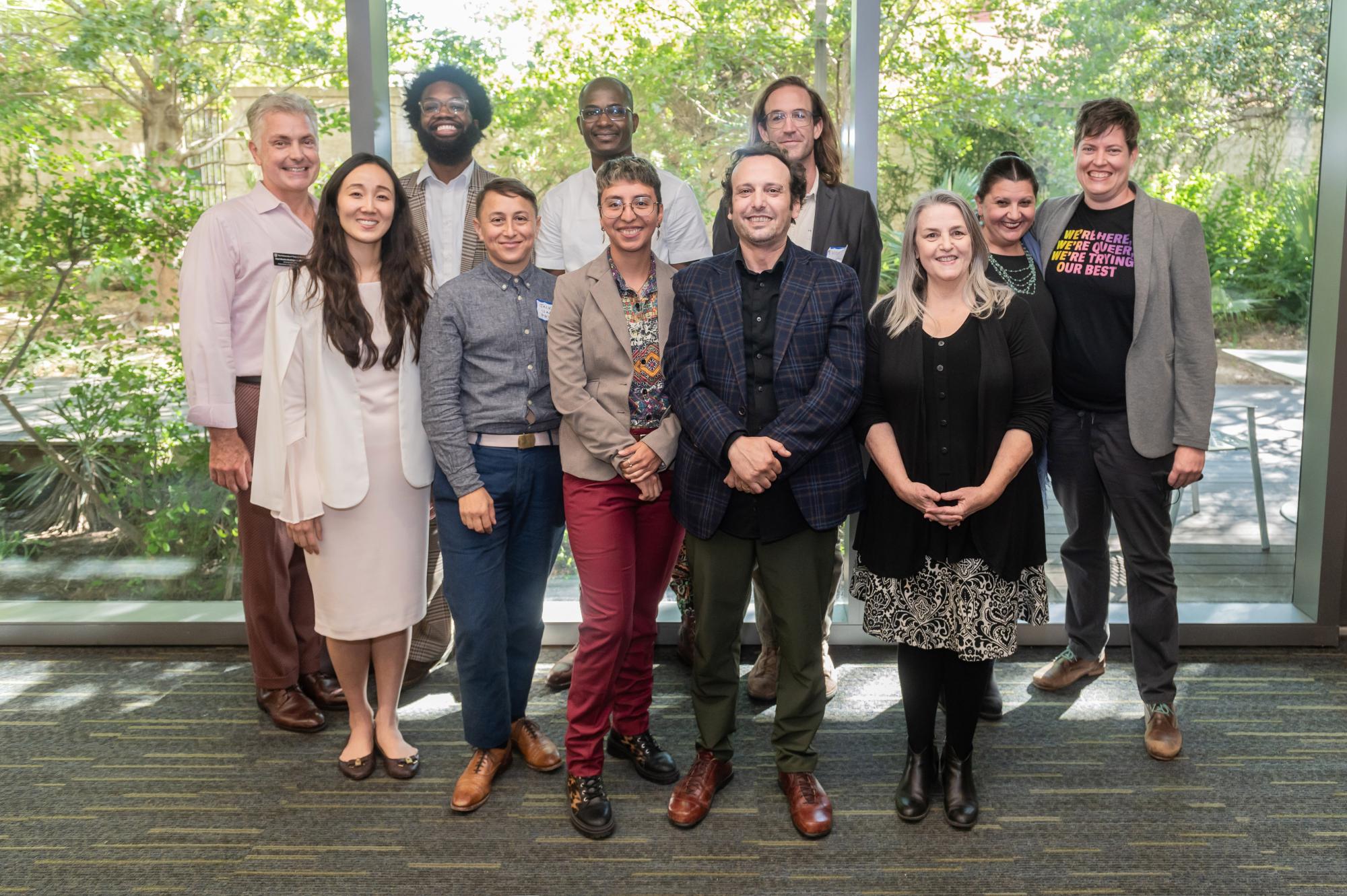 A group of 11 PPP alumni and faculty smile together, standing in front of large windows with trees behind them