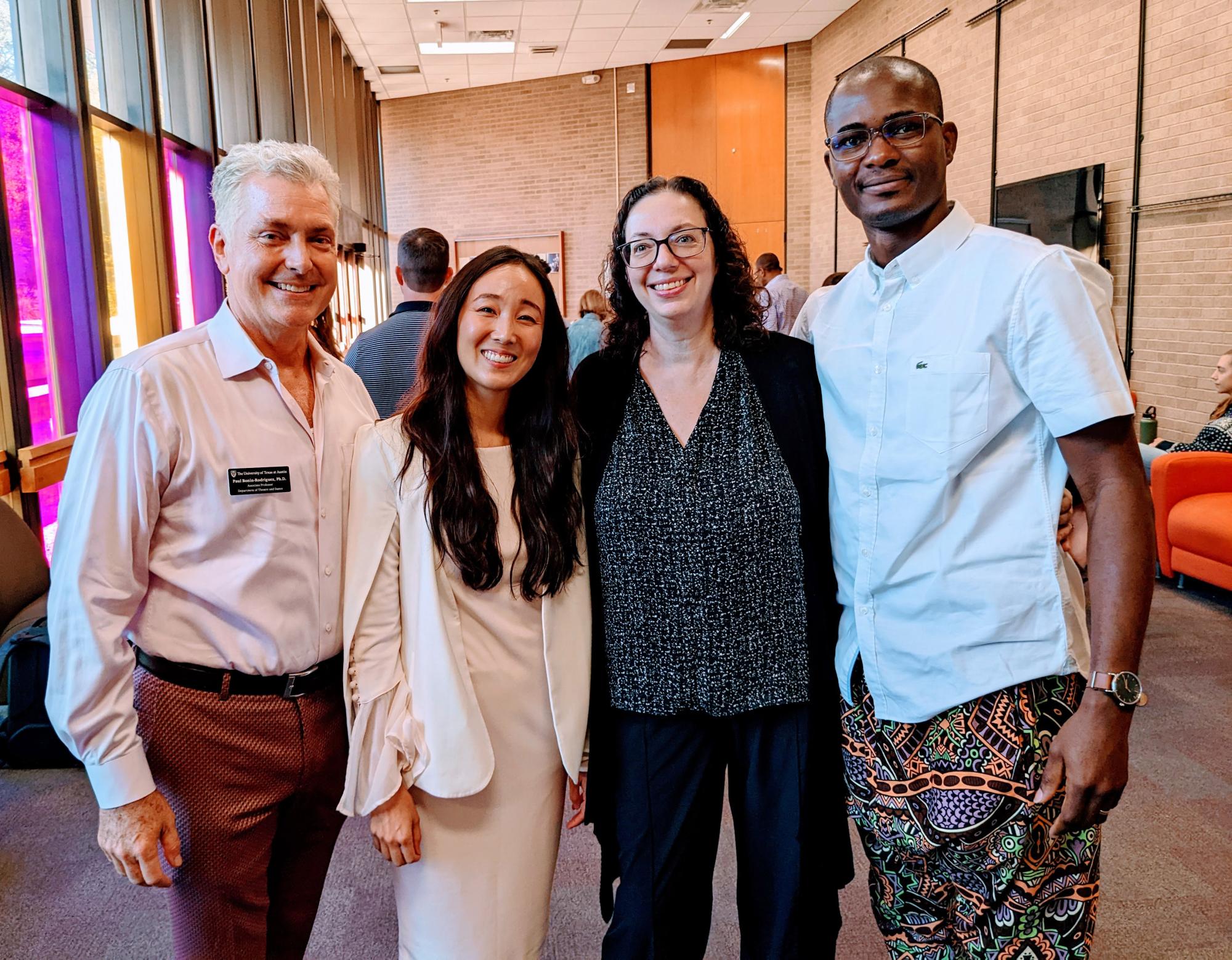 Two PPP faculty and two alumni smile and pose together in the Payne Lobby, with colorful, tinted windows behind them