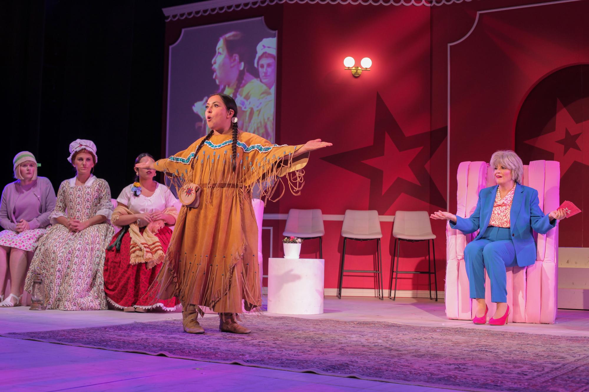 An actor walks across the stage, speaking confidently with their arms outstretched, while three other actors watch curiously and a fifth actor shrugs