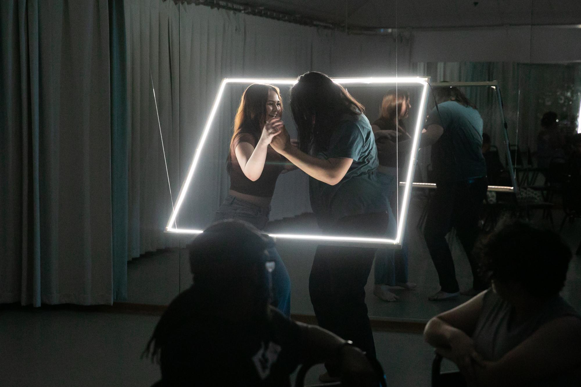 Two actors slow dance and smile at each other with a neon square hung diagonally around them