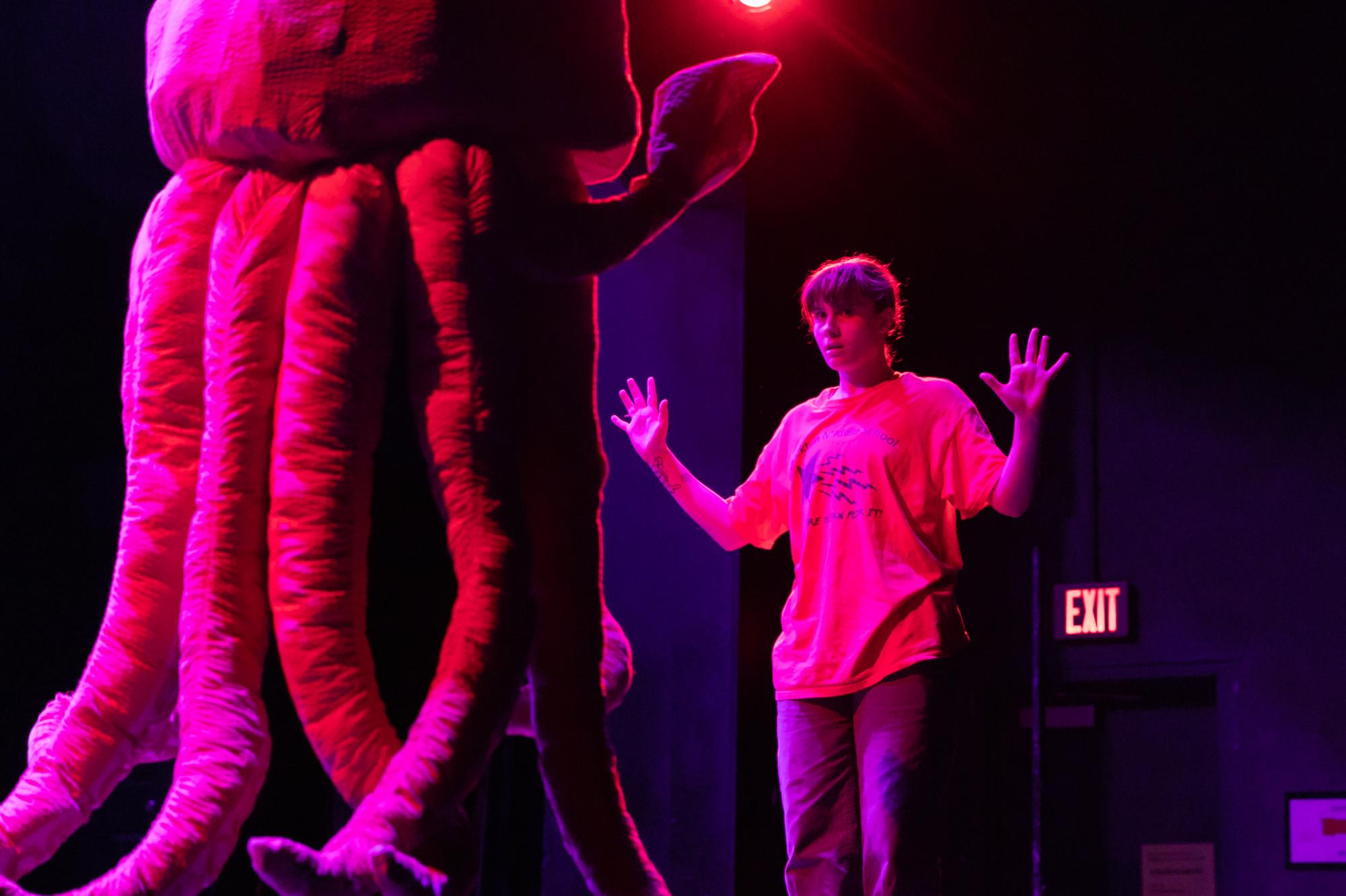 An actor holds their hands out next to them, with an actor in a pink squid costume facing them with their back to the camera