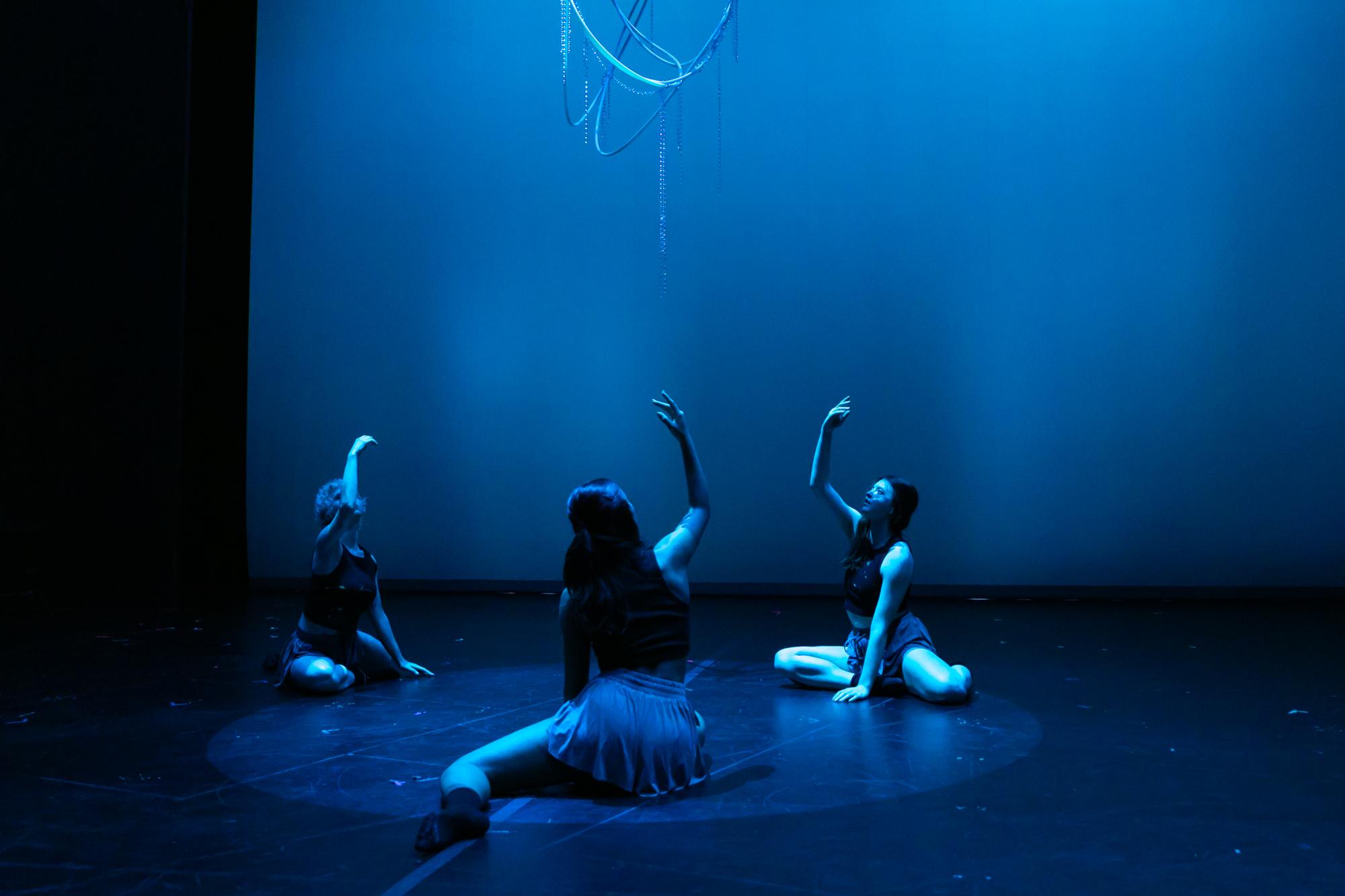three dancers sit on the Brockett Theatre floor, reaching their right arms up, washed in blue light