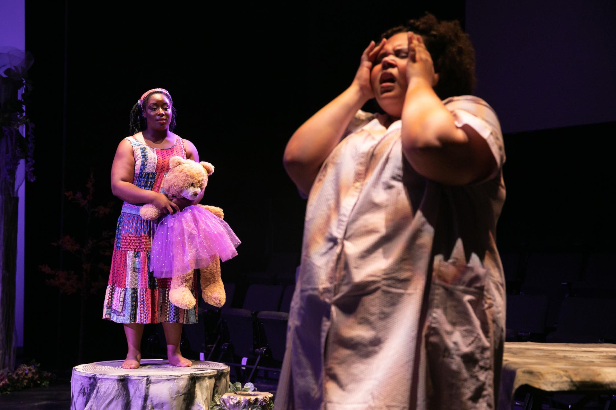 An actor wearing a long purple night gown holds their hands to their temples, while another actor in the background holding a large teddy bear watches and comforts them