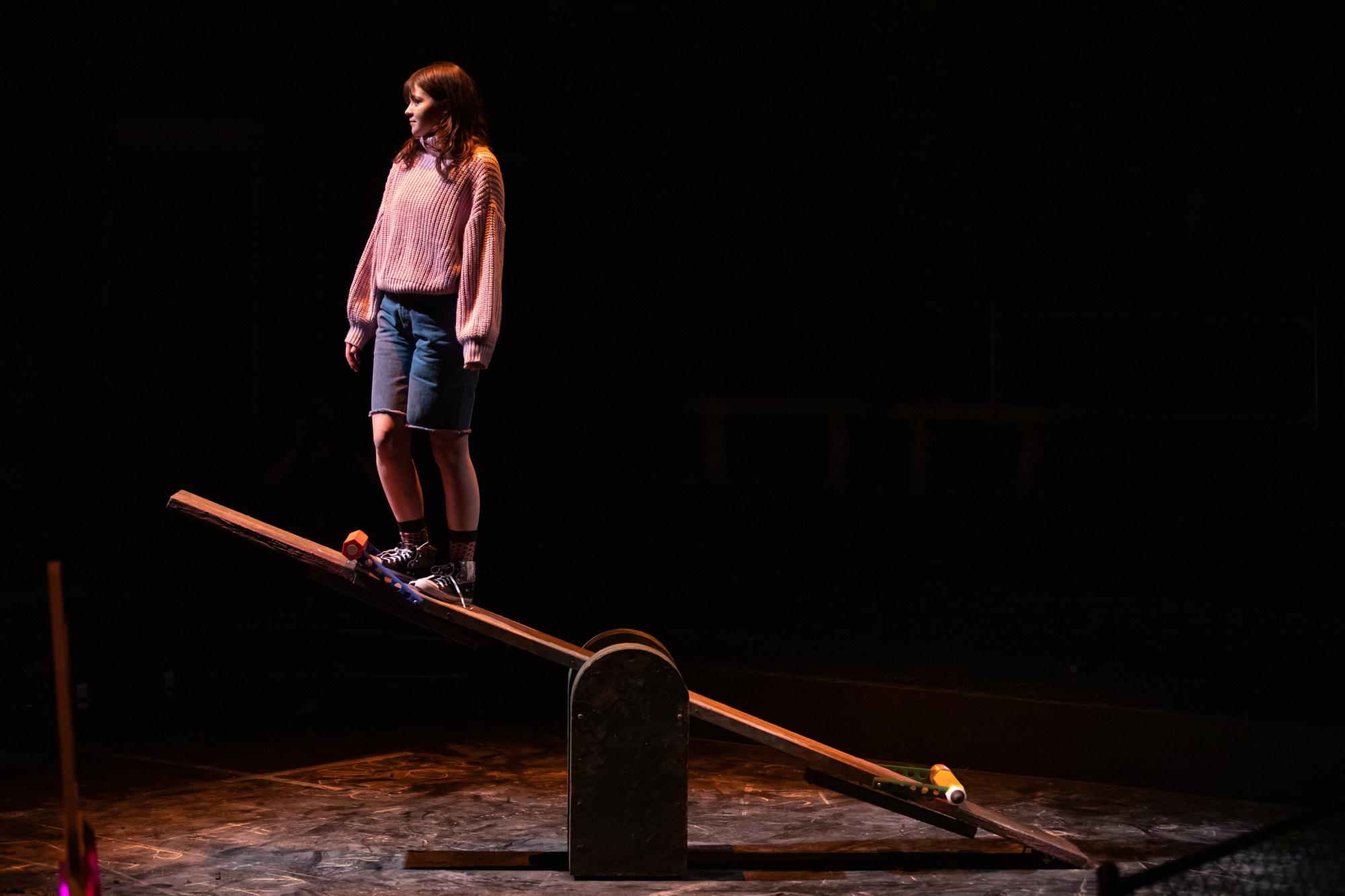 Actress Penny Lou Zimmerman as Clara stands above the ground at the end of a tall see-saw.