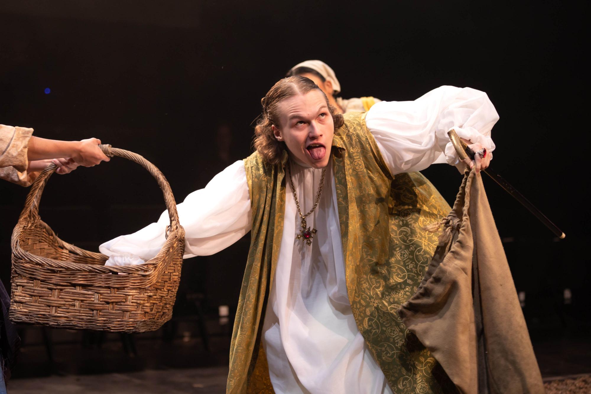 Joe Adkins as Father reaches in the basket of Frau Thera, played by Carlise Rosa, as Mia Ngyuen as Frau Sevallia looks on. His tongue is out and in his other hand is a cane and the official potato collecting bag.