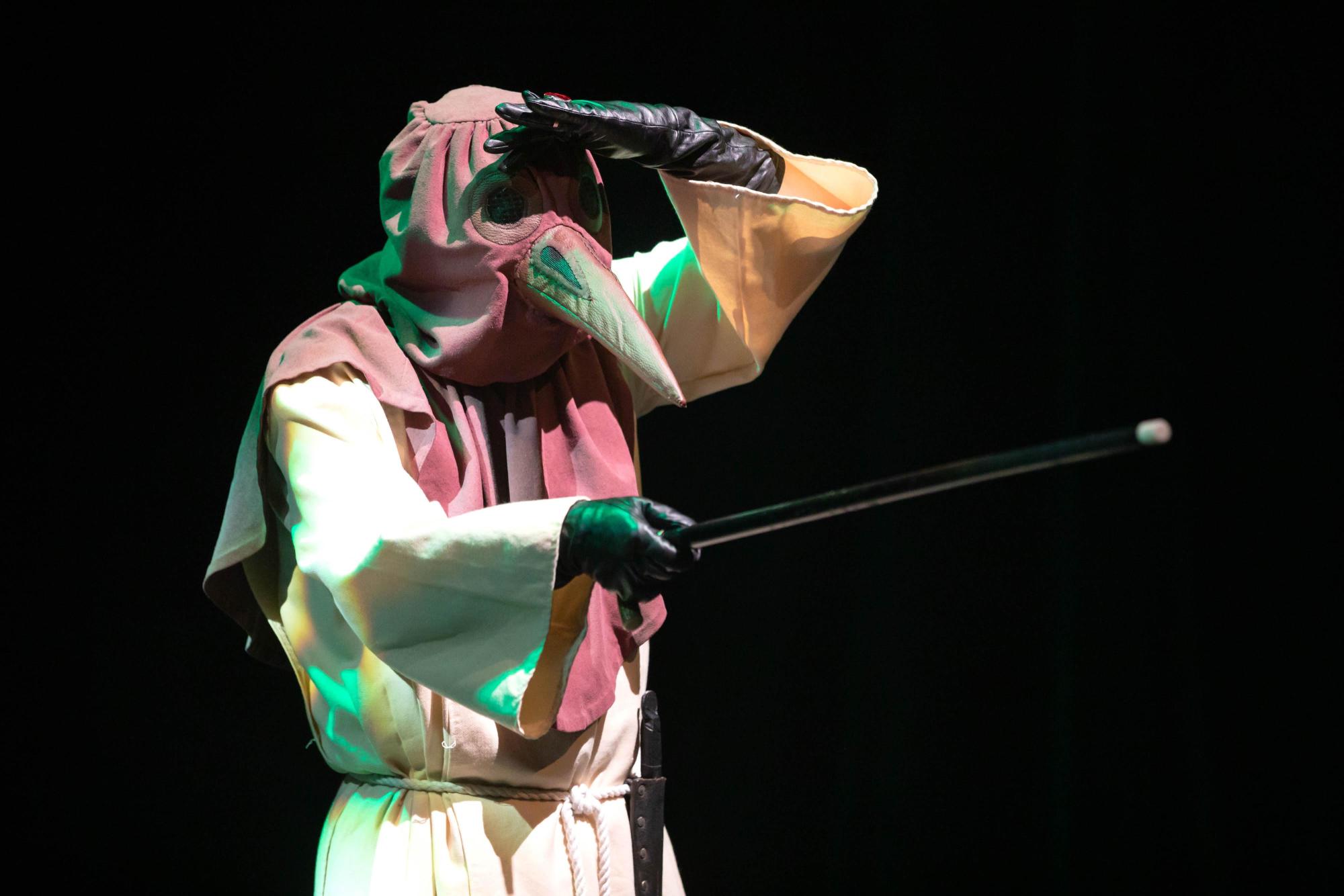 Joe Adkins as the Doctor investigates, from a distance, Frau Mika for any afflictions. He is wearing a plague mask with a beak on it.