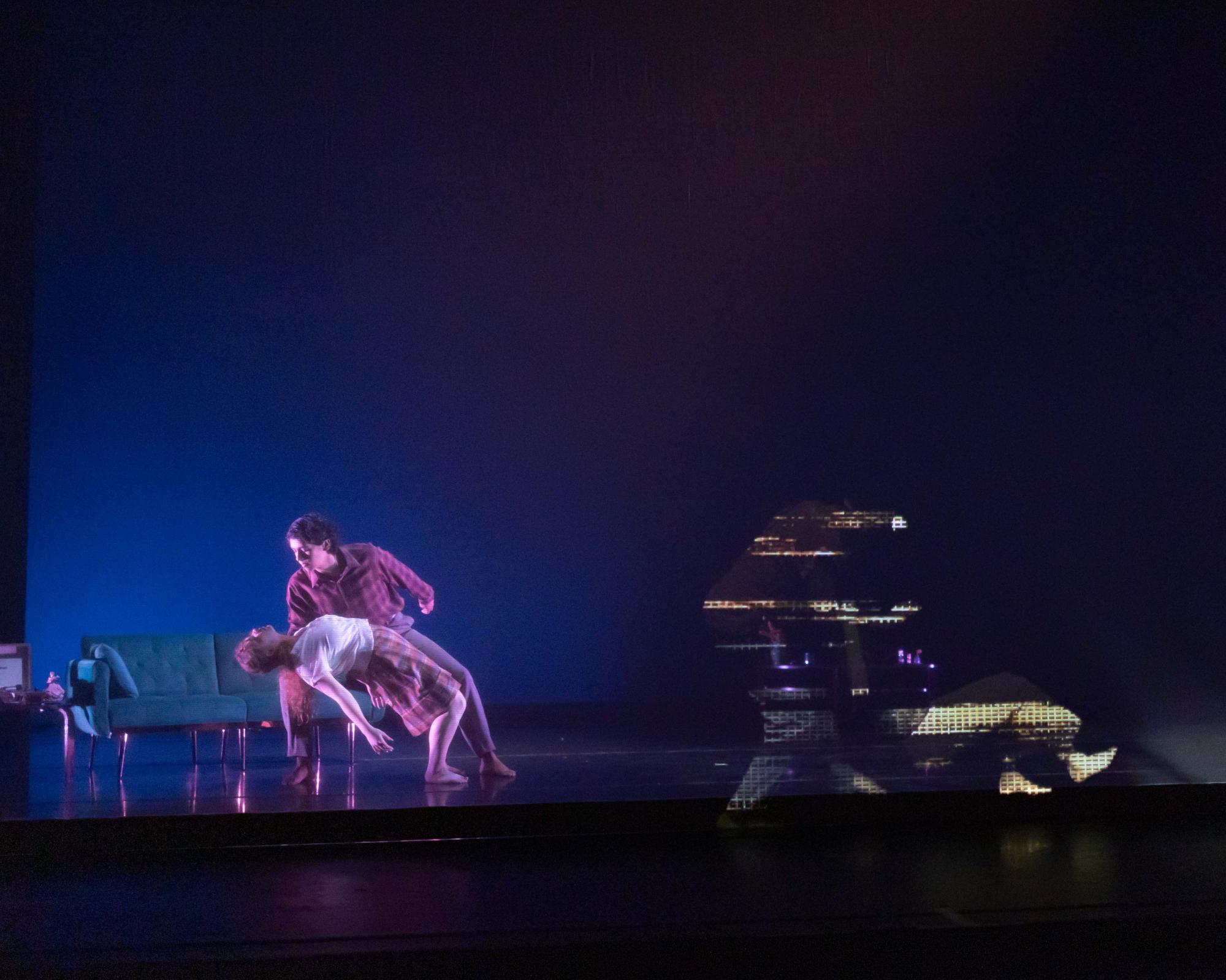 2 dancers perform a duet behind a scrim on stage. They are joined by 2 dancers that are being projected onto the stage.