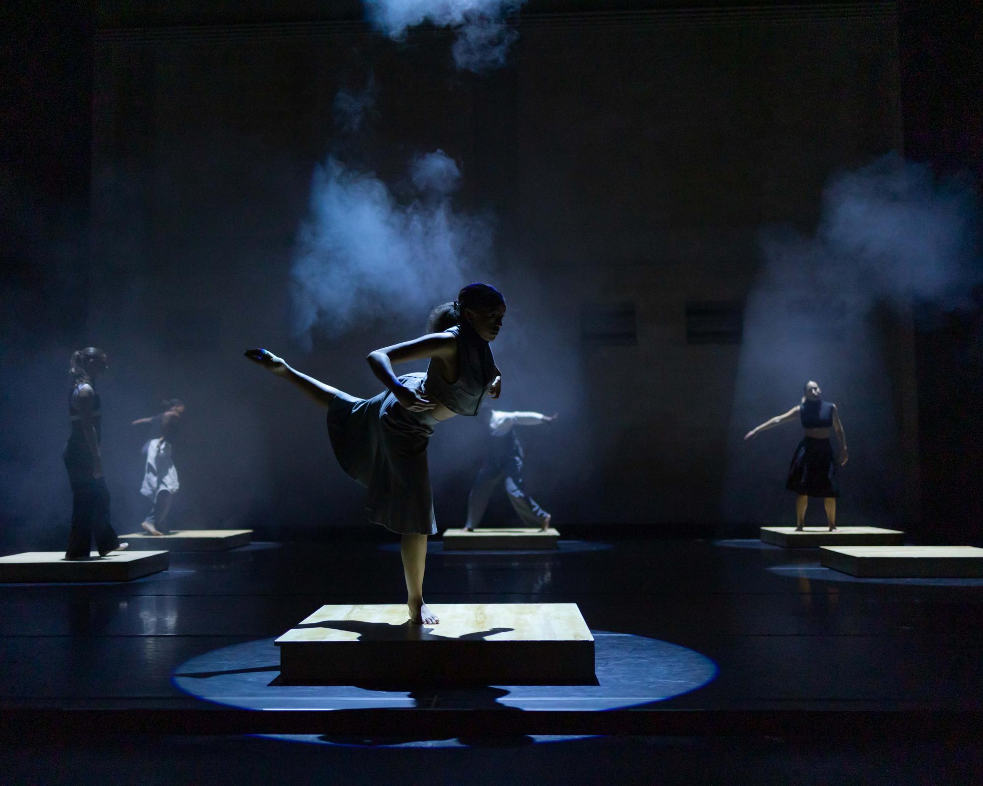 On wooden platforms stand multiple dancers scattered around the stage. A haze and dim spotlights engulf them as they move through poses.