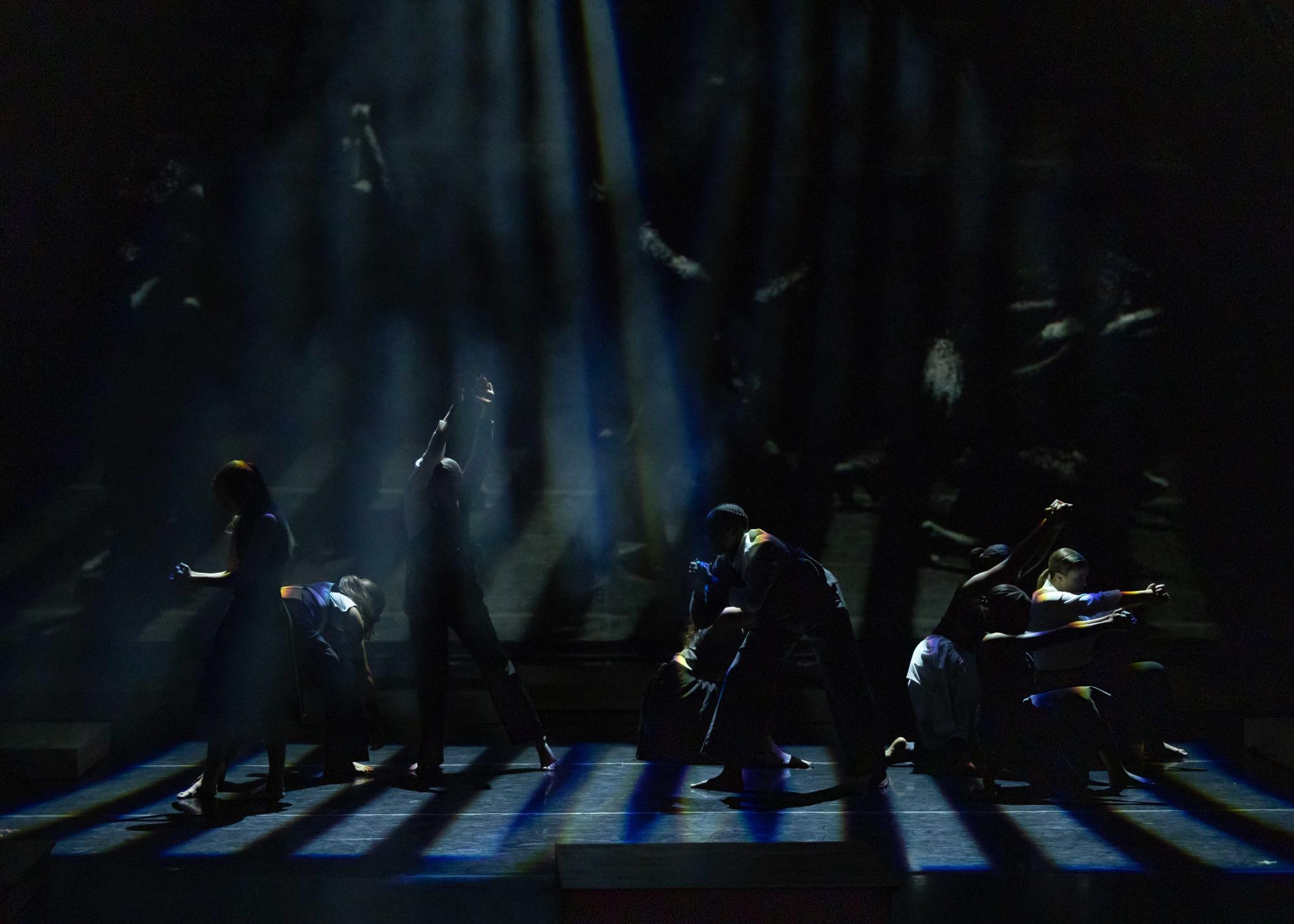 Light that is being refracted into rainbows shines down on a group of dancers in dynamic poses. The stage is mostly dark except for these beams of light that barely illuminate the dancers.