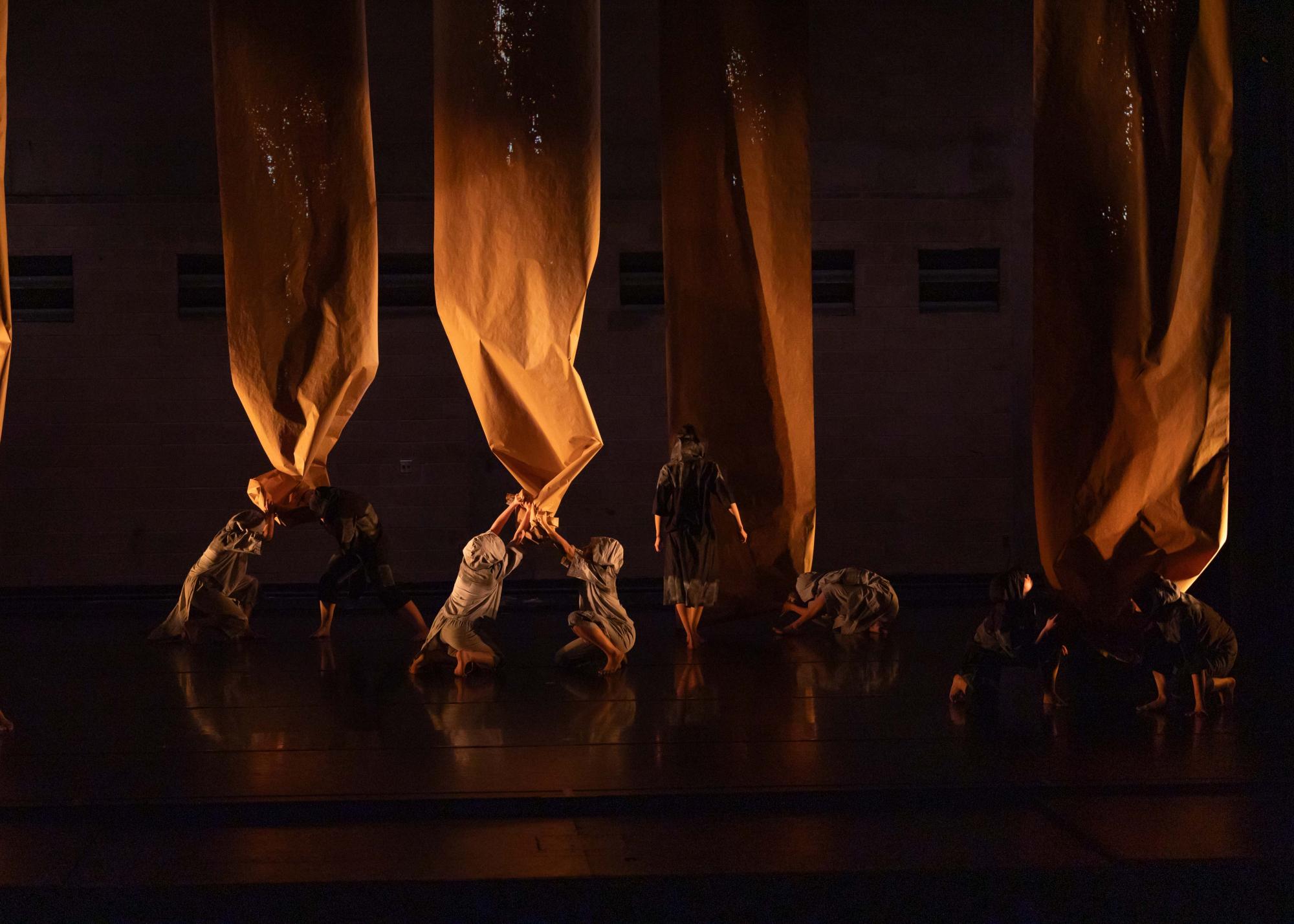 Dancers cloaked in grey and black crouch at the bottom of strips of long brown paper dangling from the ceiling. They crumple it, ruin it. They are the rust of the world eating away at the good.