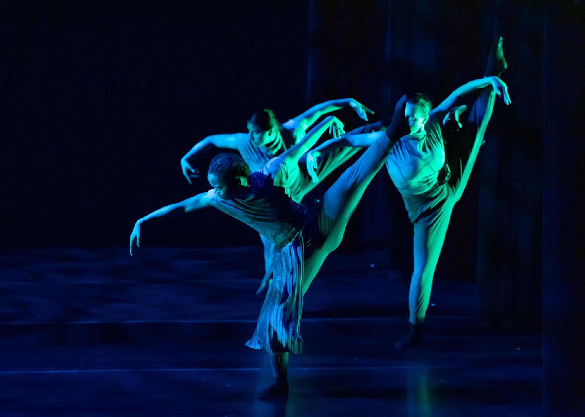 Three dancers balance on their right legs, their arms outstretched beside them and their left legs pointed diagonally behind them