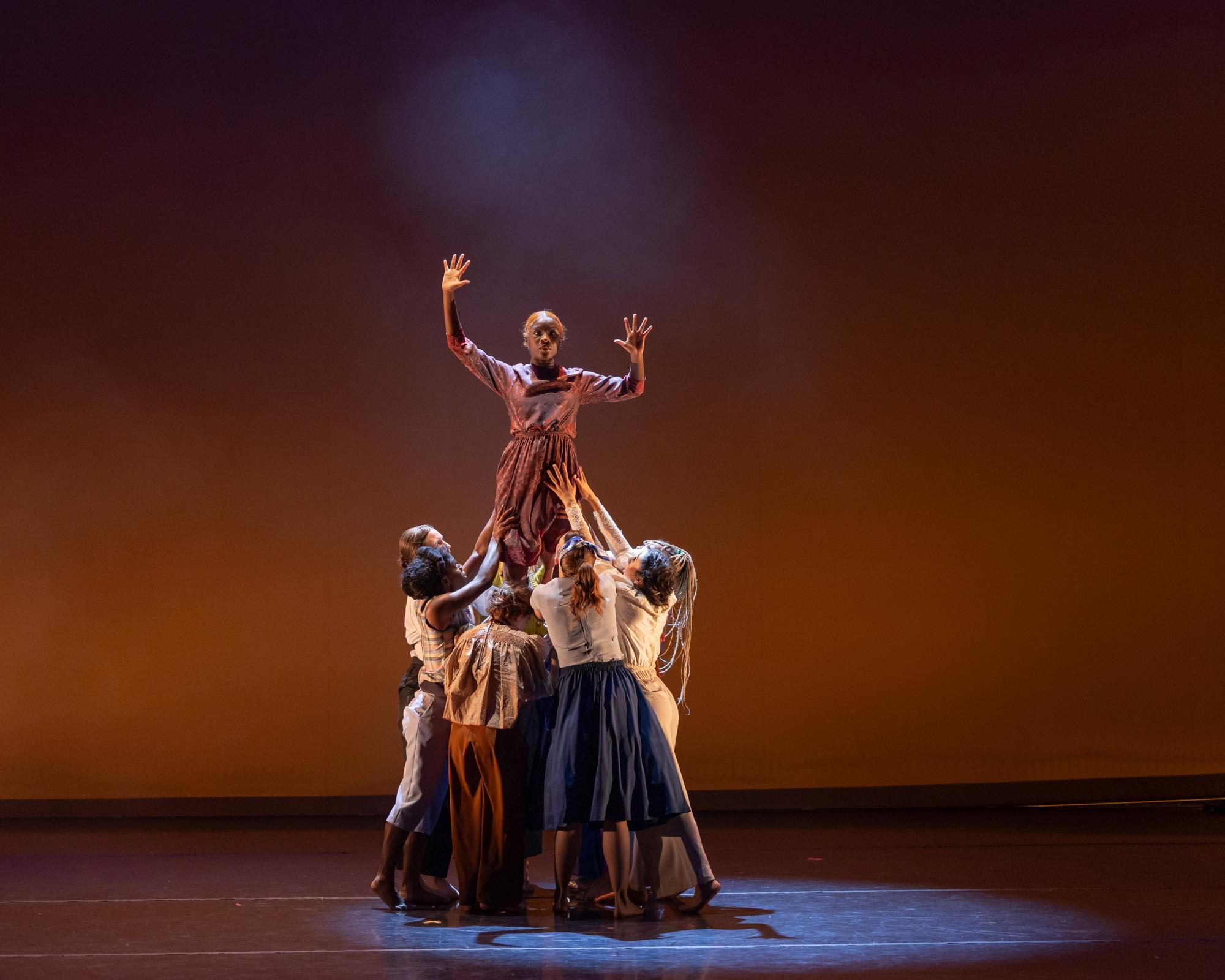 A group of at least seven dancers hold up another dancer, who is holding her arms out at right angles next to her