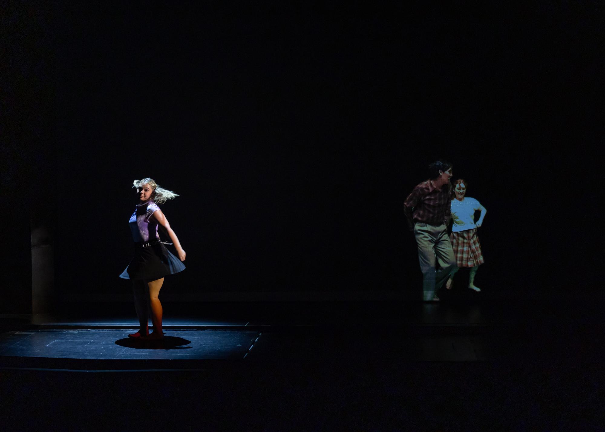  A dancer twists in a spotlight on stage, with two other dancers projected on a scrim next to her