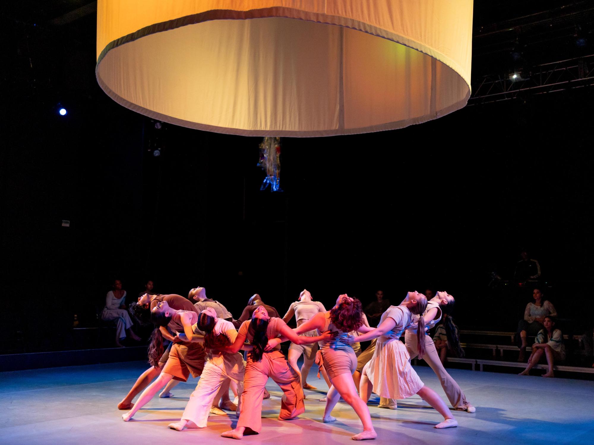 Dancers in neutral outfits stand in circle with one leg extended behind them. They are watching their coats float above their heads.