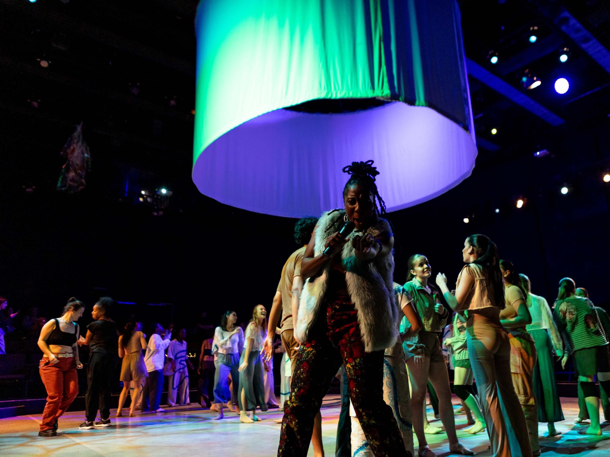 MC, Aysha Upchurch, points at the camera while dancers move behind her.