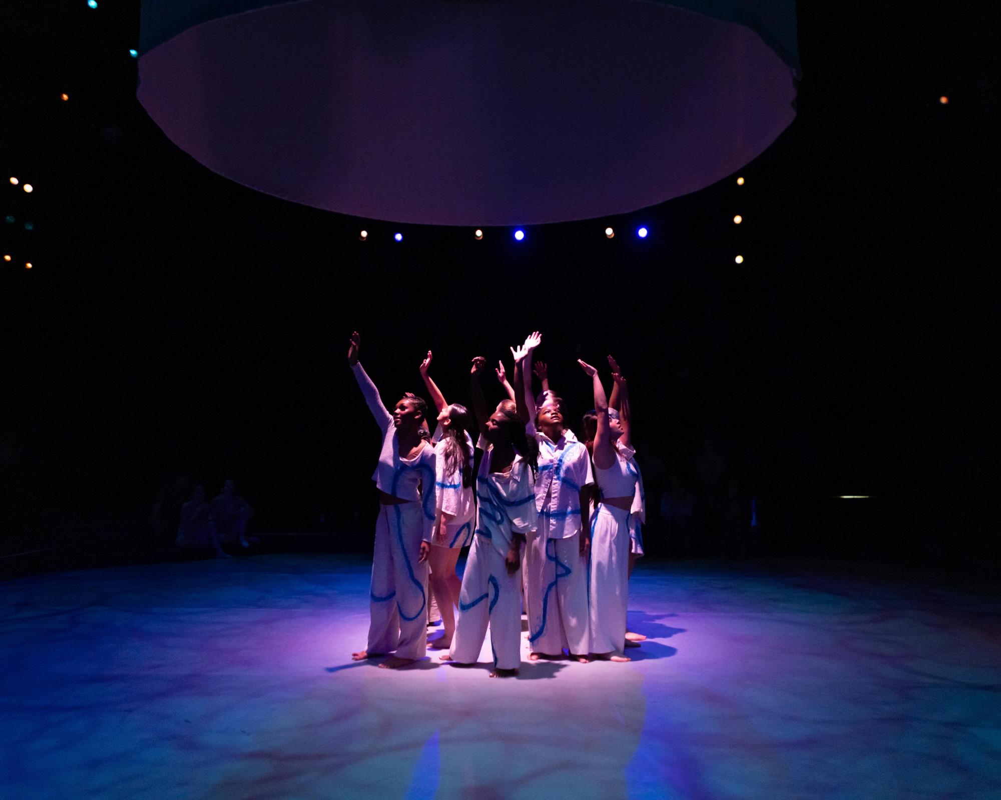 Dancers in white outfits with blue embroidery stand in a circle with an arm raised to the sky. They face the audience. They are saying goodbye.