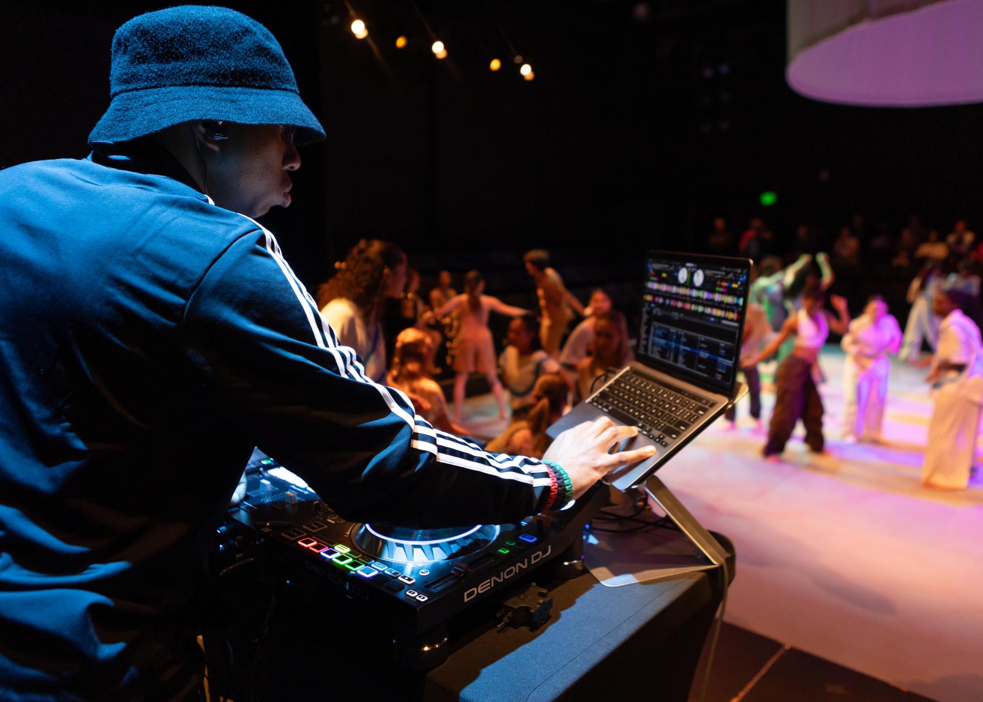 DJ, Earnest Mazique, mixes music in the foreground of the image while dancers can be seen in the background over his shoulder.