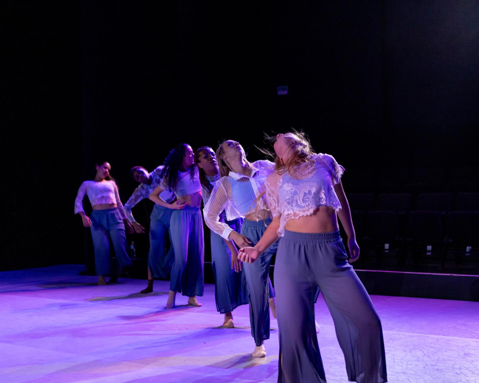 Dancers stand in a diagonal basked in purple light, they writhe with emotion.