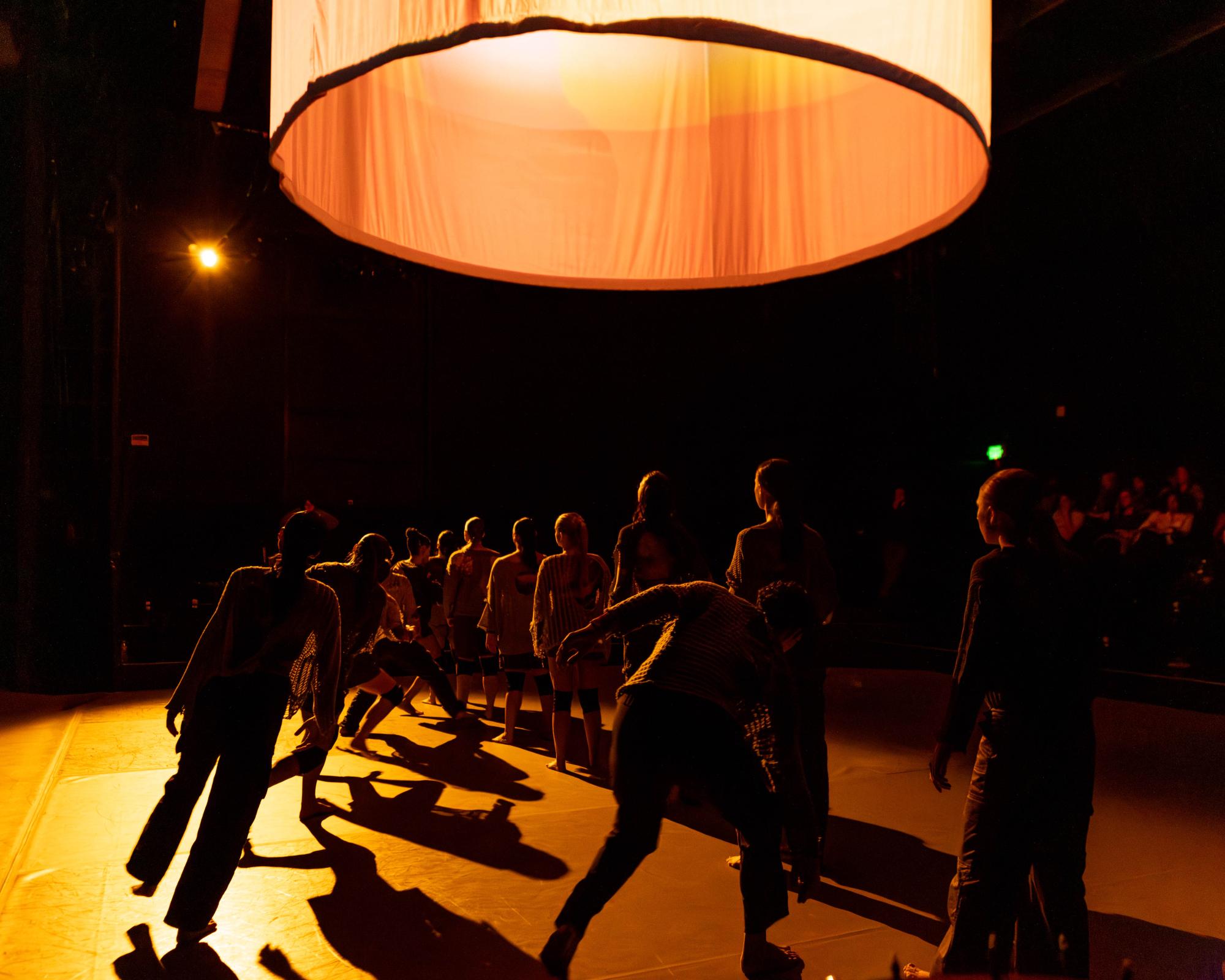 Cloaked in darkness, standing against a dimmed light, dancers are lined up as some move in and out of the diagonal they are standing in.
