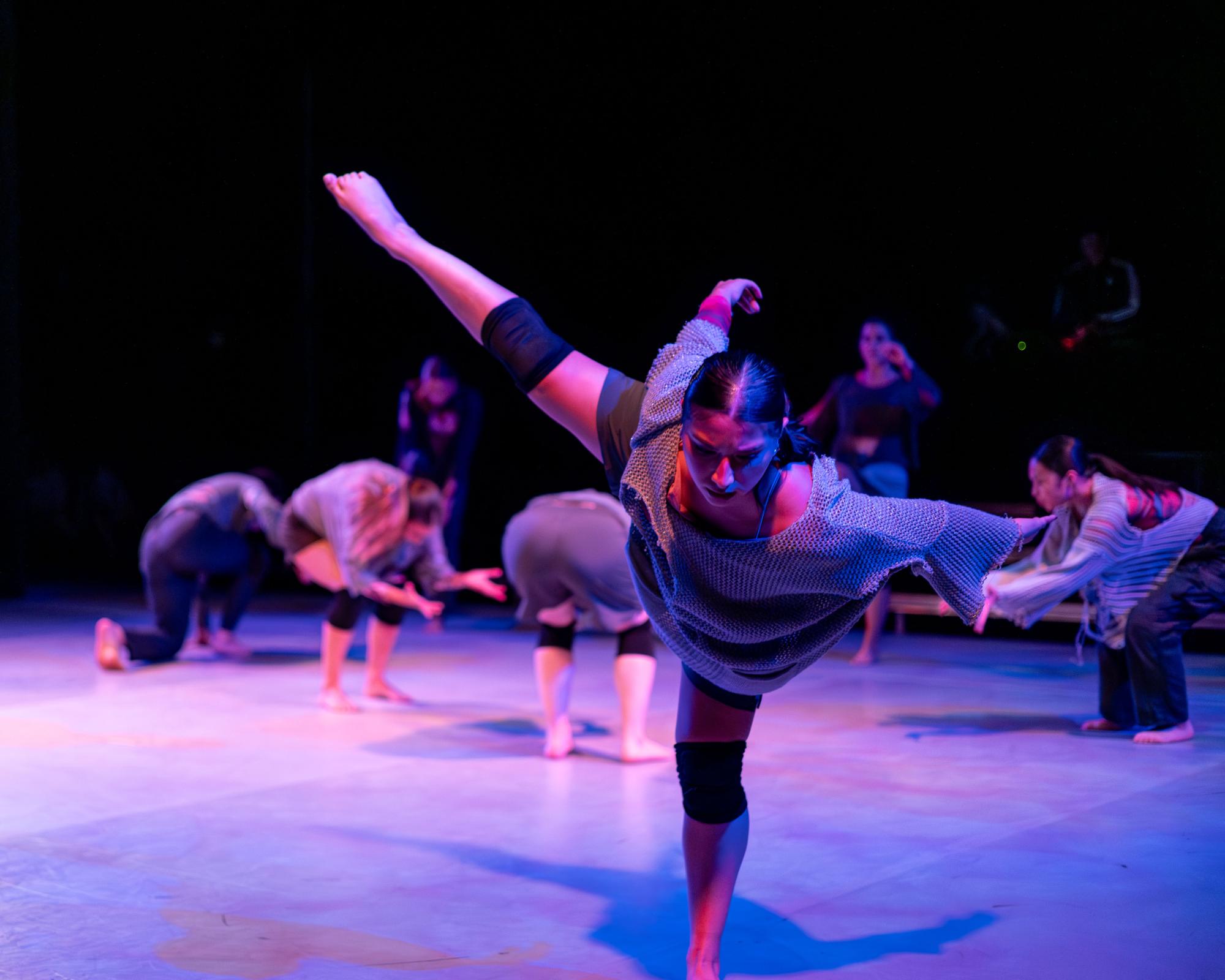 Amid a crowd of dancers crouched down, one dancer stands against the crowd crouching with their leg extended toward the side.