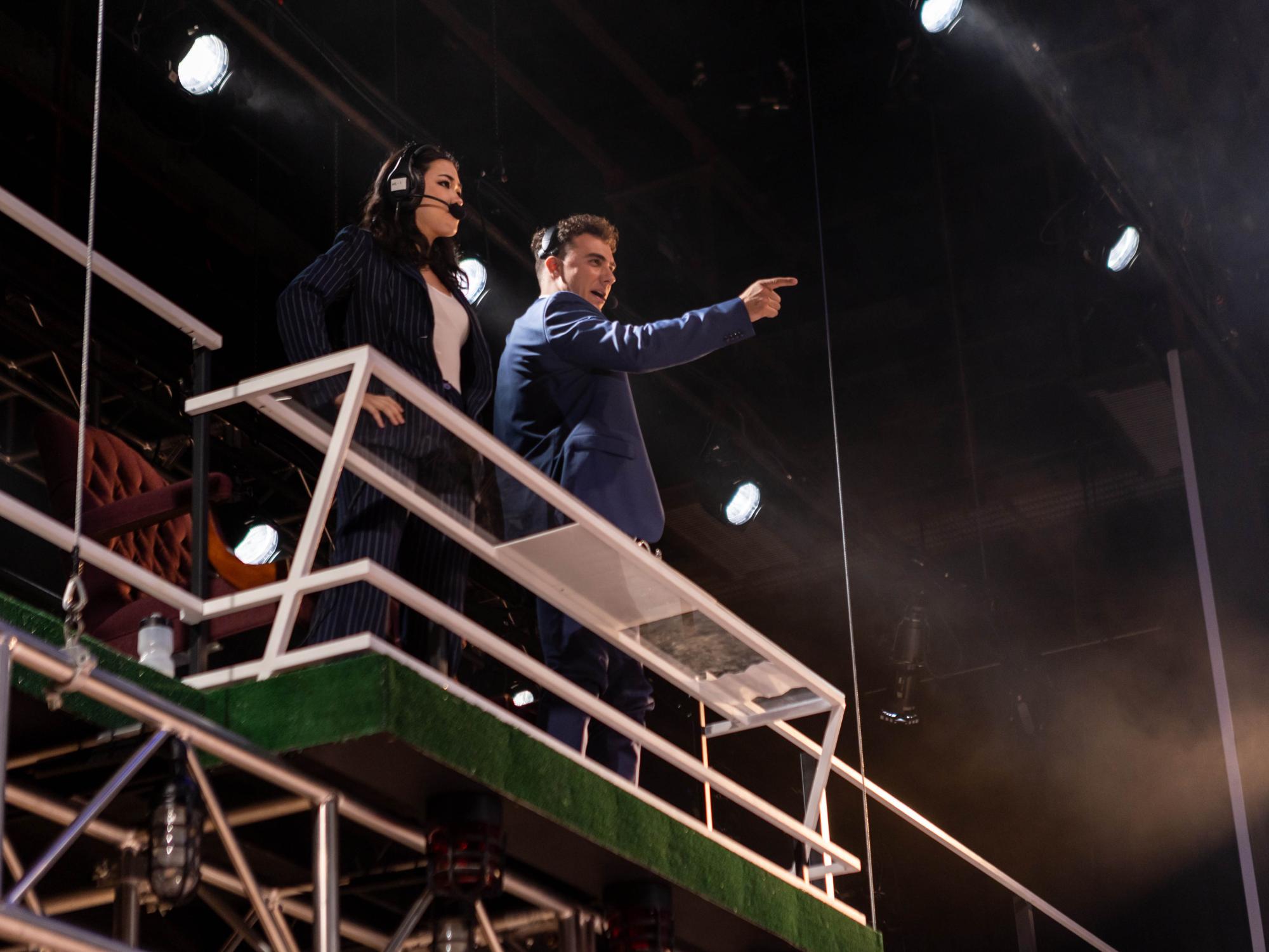 The two announcers played by Natalie Tran and Jake Wilson stand on their perch pointing and presenting a power pose, overlooking the action at Wemberly Kitchen