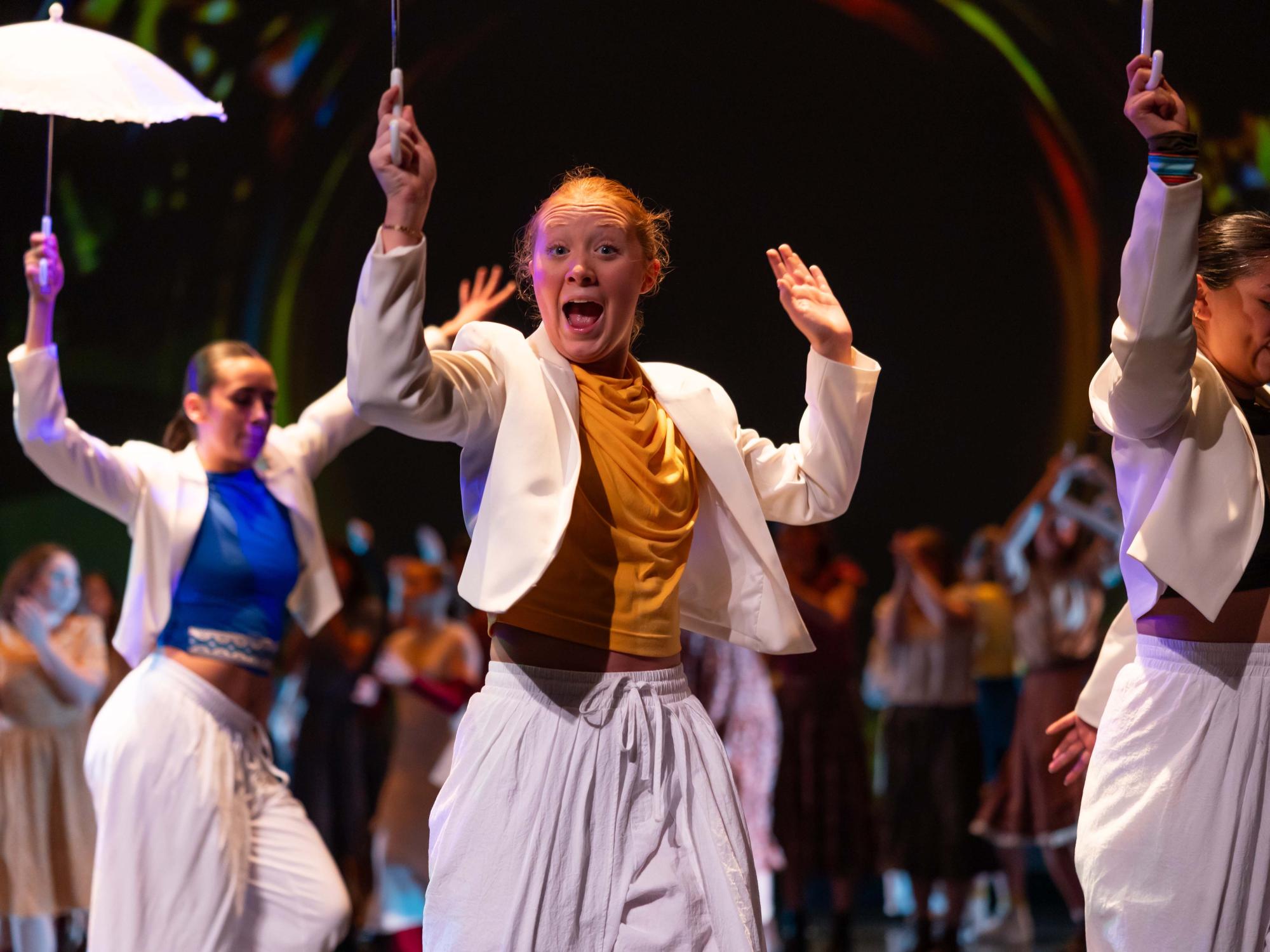In The Altar of Humanity The Convocation The Resolution The Benediction, a dancer in white and yellow smiles brightly at the camera, celebrating.
