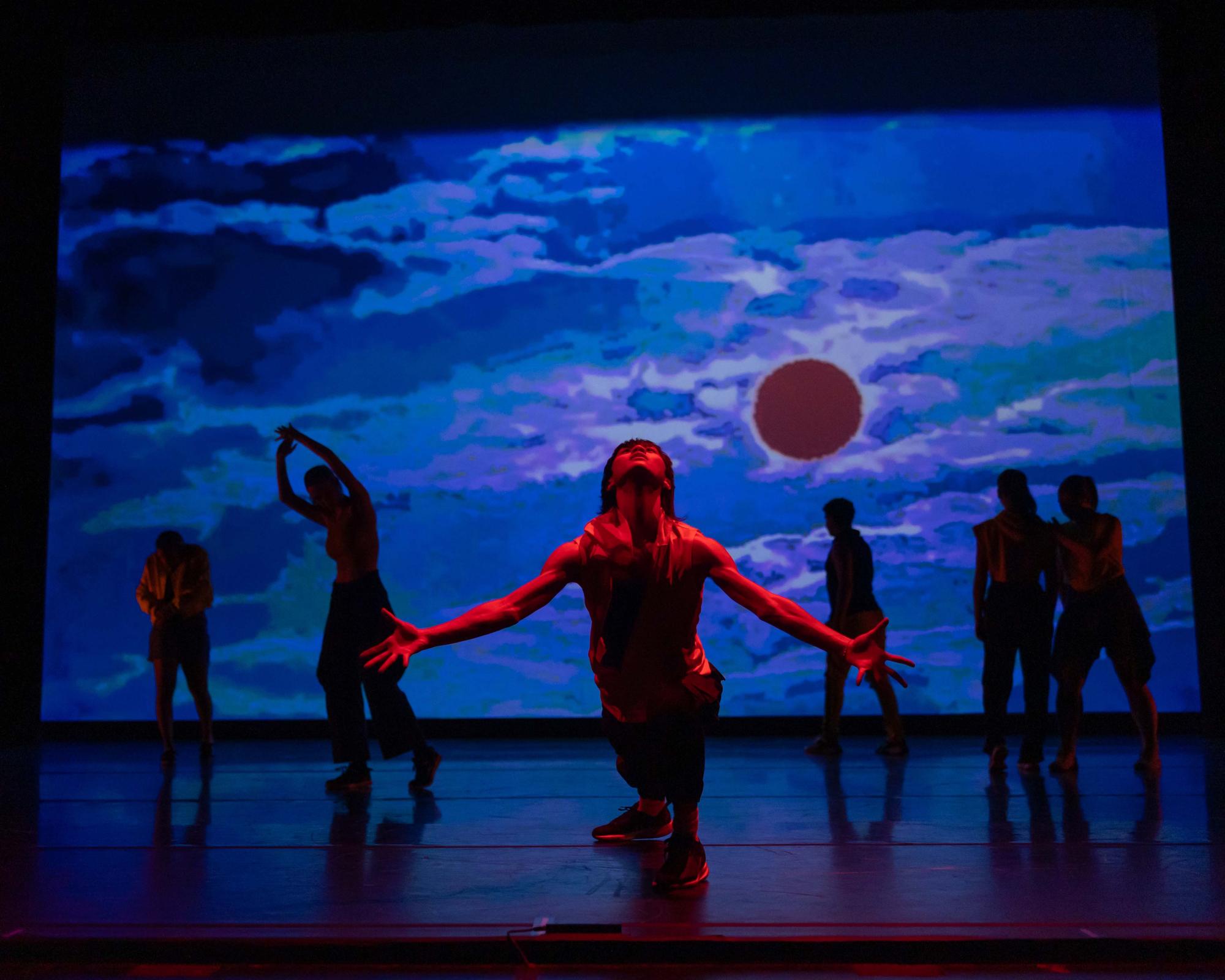 A dancer in Lone. Lean. Less. reaches their arms out to the audience while a red sun is alight behind them.